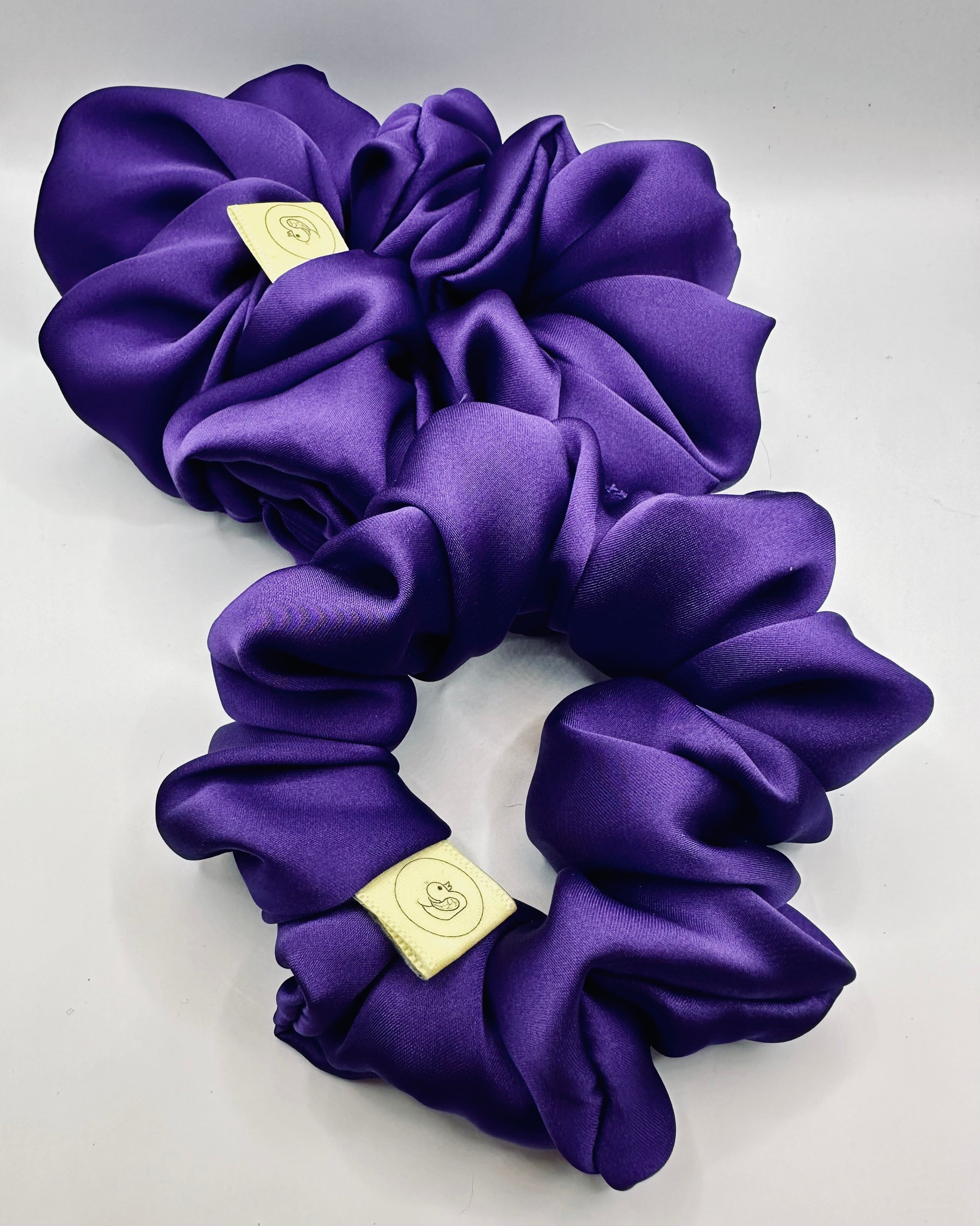 This shows a luxurious scrunchie made of silky textured fabric in a deep purple colour giving it an elegant and slightly reflective appearance.The scrunchie is gathered and voluminous creating soft folds and a plush look.A small yellow label tag with a picture of a duck is attached to the scrunchie adding a touch of branding.The overall aesthetic is soft, elegant and stylish. Perfect for those with sensitive scalp or hair loss