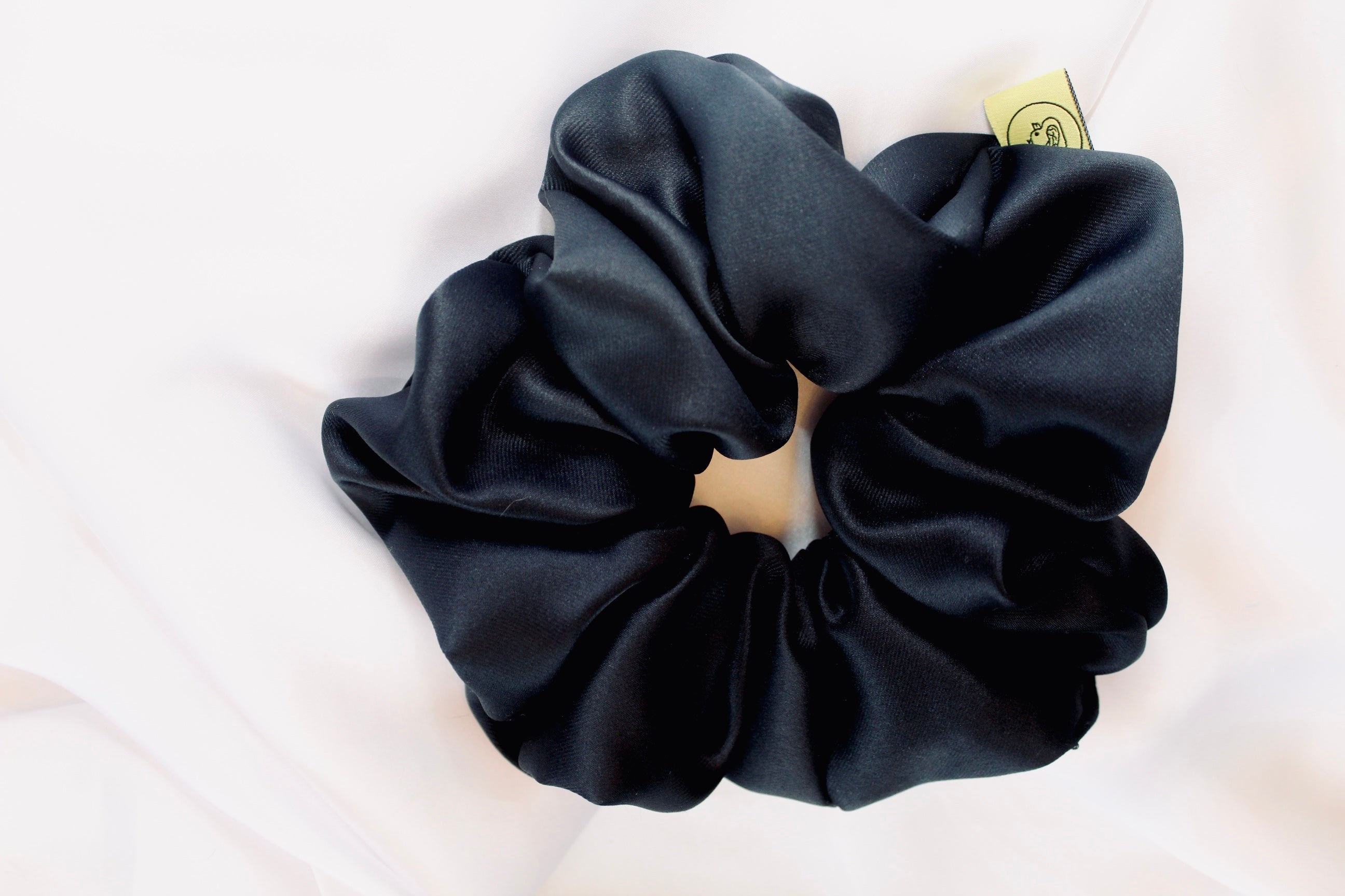 This shows a luxurious scrunchie made of silky textured fabric in a black thick fabric giving it an elegant and slightly reflective appearance.The scrunchie is gathered and voluminous creating soft folds and a plush look.A small yellow label tag with a picture of a duck is attached to the scrunchie adding a touch of branding.The overall aesthetic is soft, elegant and stylish. Perfect for those with sensitive scalp or hair loss