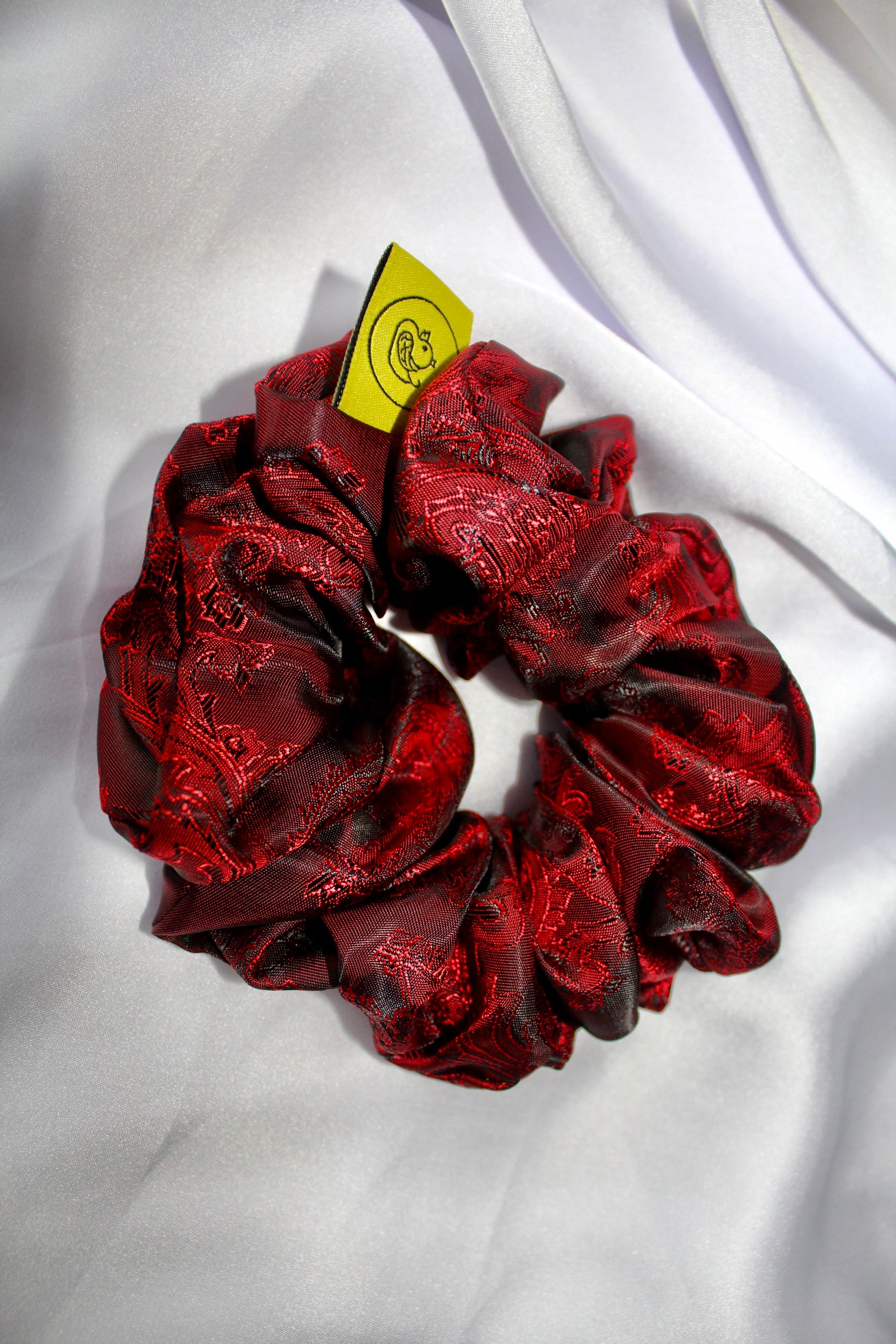 This shows a luxurious scrunchie made of silky textured fabric in a red and black colour.The fabric has a subtle paisley pattern wove into it, giving it an elegant and slightly reflective appearance.The scrunchie is gathered and voluminous creating soft folds and a plush look.A small yellow label tag with a picture of a duck is attached to the scrunchie adding a touch of branding.The overall aesthetic is soft, elegant and stylish. Perfect for those with sensitive scalp or hair loss