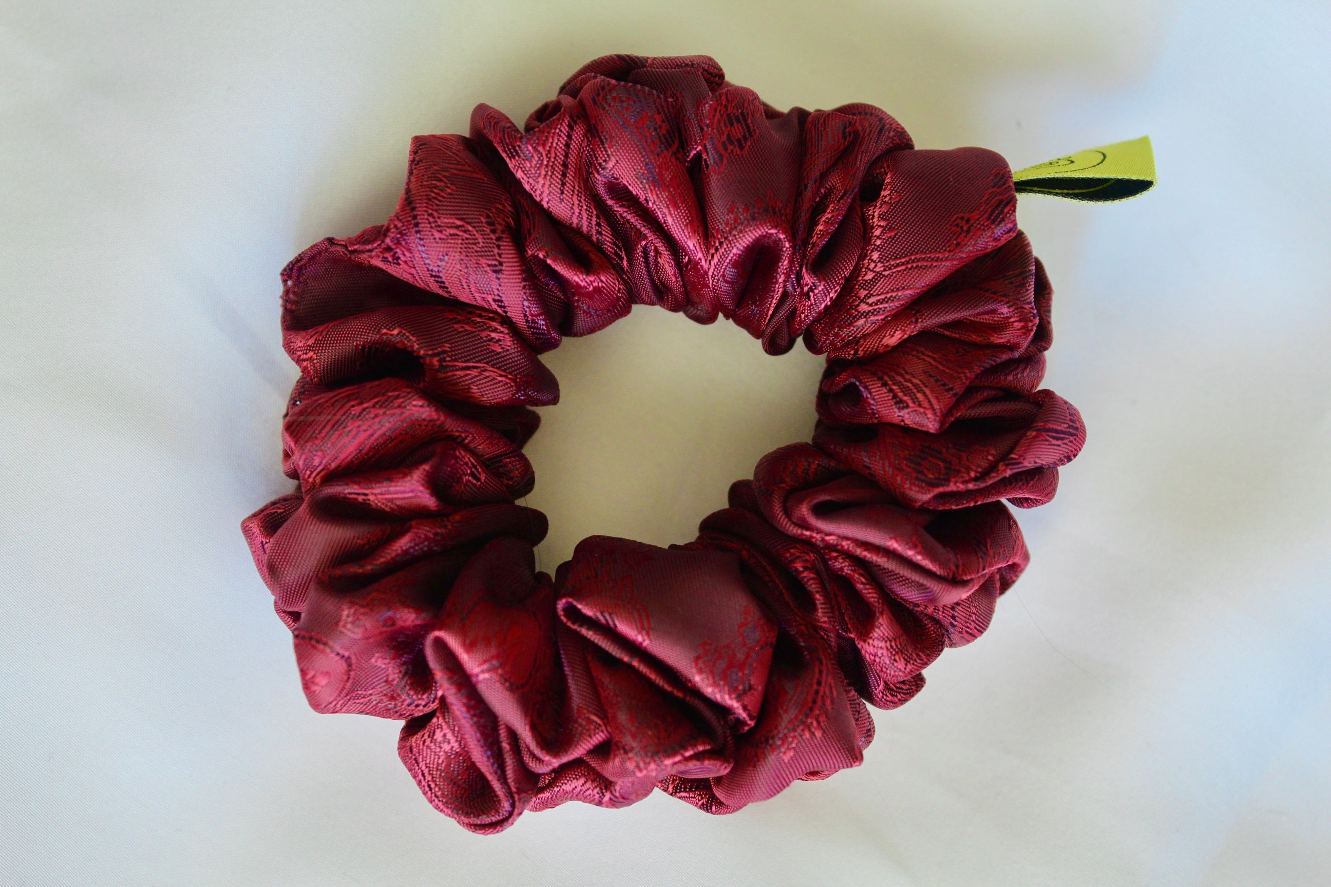 This shows a luxurious scrunchie made of silky textured fabric in a brown mocha and gold.The fabric has a subtle paisley pattern wove into it, giving it an elegant and slightly reflective appearance.The scrunchie is gathered and voluminous creating soft folds and a plush look.A small yellow label tag with a picture of a duck is attached to the scrunchie adding a touch of branding.The overall aesthetic is soft, elegant and stylish. Perfect for those with sensitive scalp or hair loss