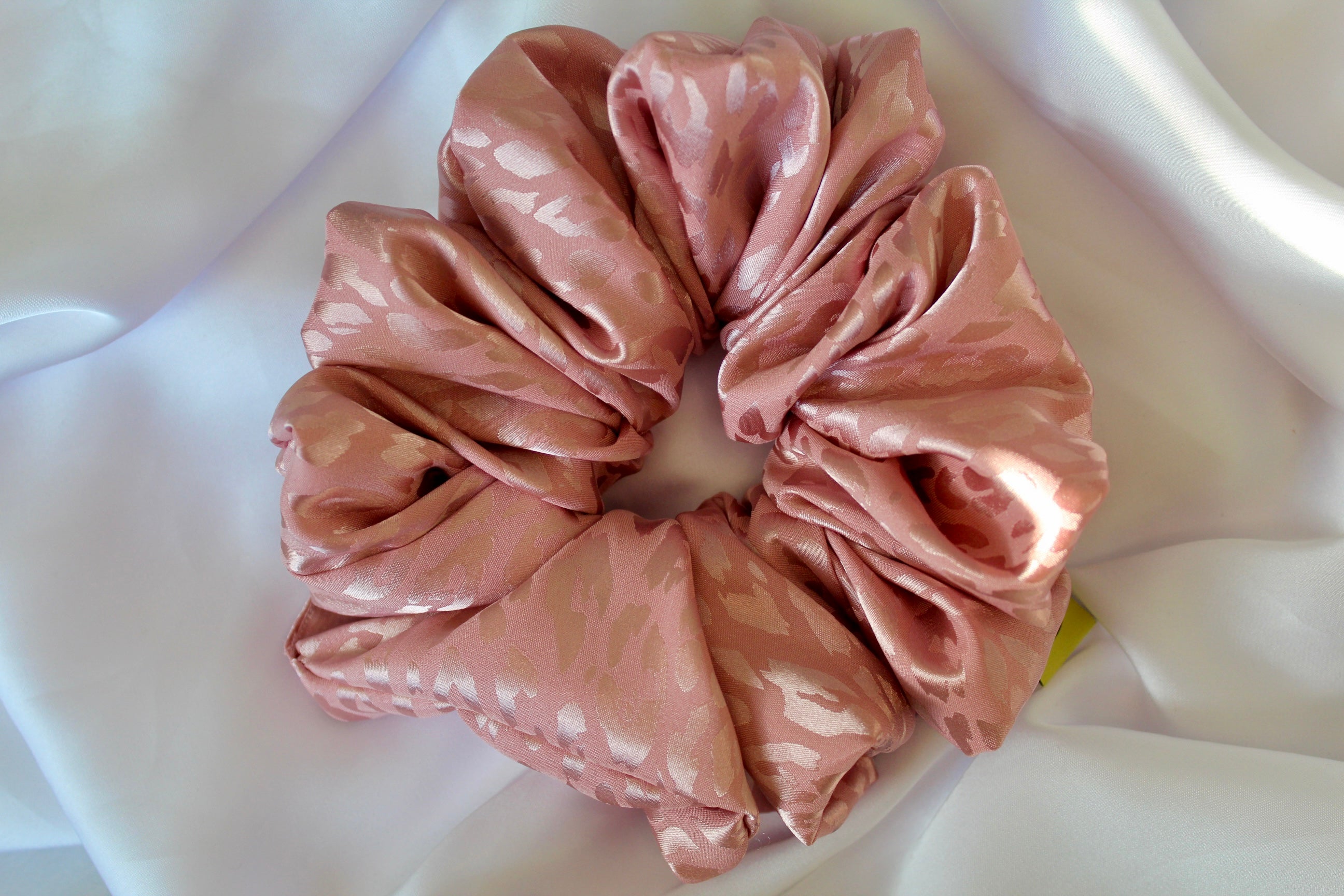 This shows a luxurious scrunchie made of silky textured fabric in a dusty rose pink colour.The fabric has a subtle leopard print pattern wove into it, giving it an elegant and slightly reflective appearance.The scrunchie is gathered and voluminous creating soft folds and a plush look.A small yellow label tag with a picture of a duck is attached to the scrunchie adding a touch of branding.The overall aesthetic is soft, elegant and stylish. Perfect for those with sensitive scalp or hair loss