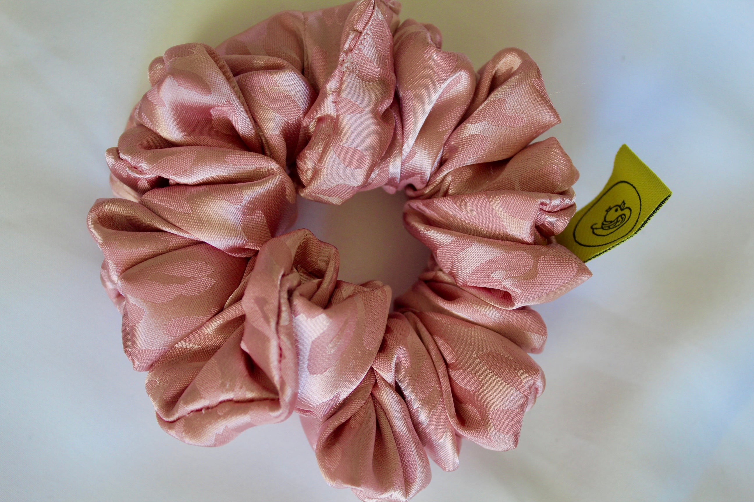 This shows a luxurious scrunchie made of silky textured fabric in a dusty rose pink colour.The fabric has a subtle leopard print pattern wove into it, giving it an elegant and slightly reflective appearance.The scrunchie is gathered and voluminous creating soft folds and a plush look.A small yellow label tag with a picture of a duck is attached to the scrunchie adding a touch of branding.The overall aesthetic is soft, elegant and stylish. Perfect for those with sensitive scalp or hair loss