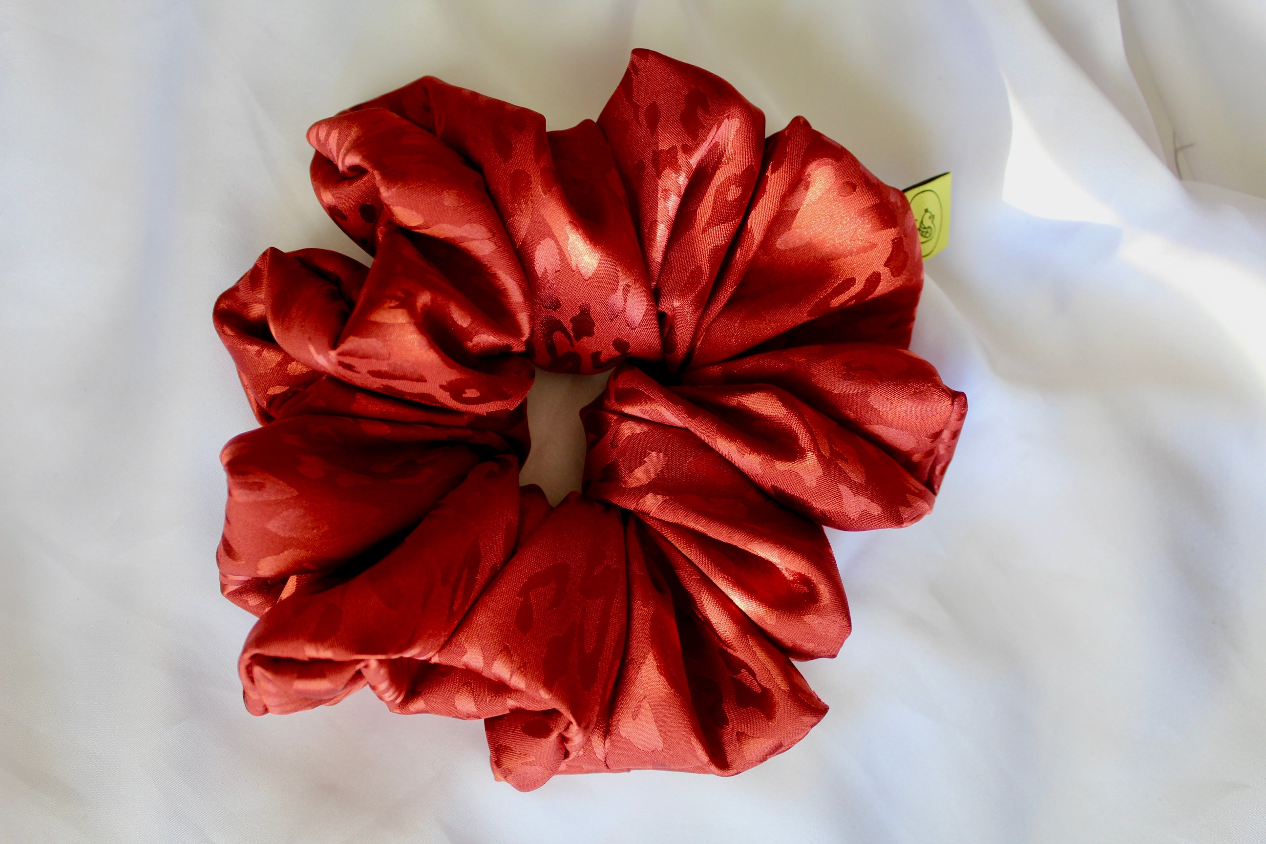 This shows a luxurious scrunchie made of silky textured fabric in a burnt orange/red colour.The fabric has a subtle leopard print pattern wove into it, giving it an elegant and slightly reflective appearance.The scrunchie is gathered and voluminous creating soft folds and a plush look.A small yellow label tag with a picture of a duck is attached to the scrunchie adding a touch of branding.The overall aesthetic is soft, elegant and stylish. Perfect for those with sensitive scalp or hair loss