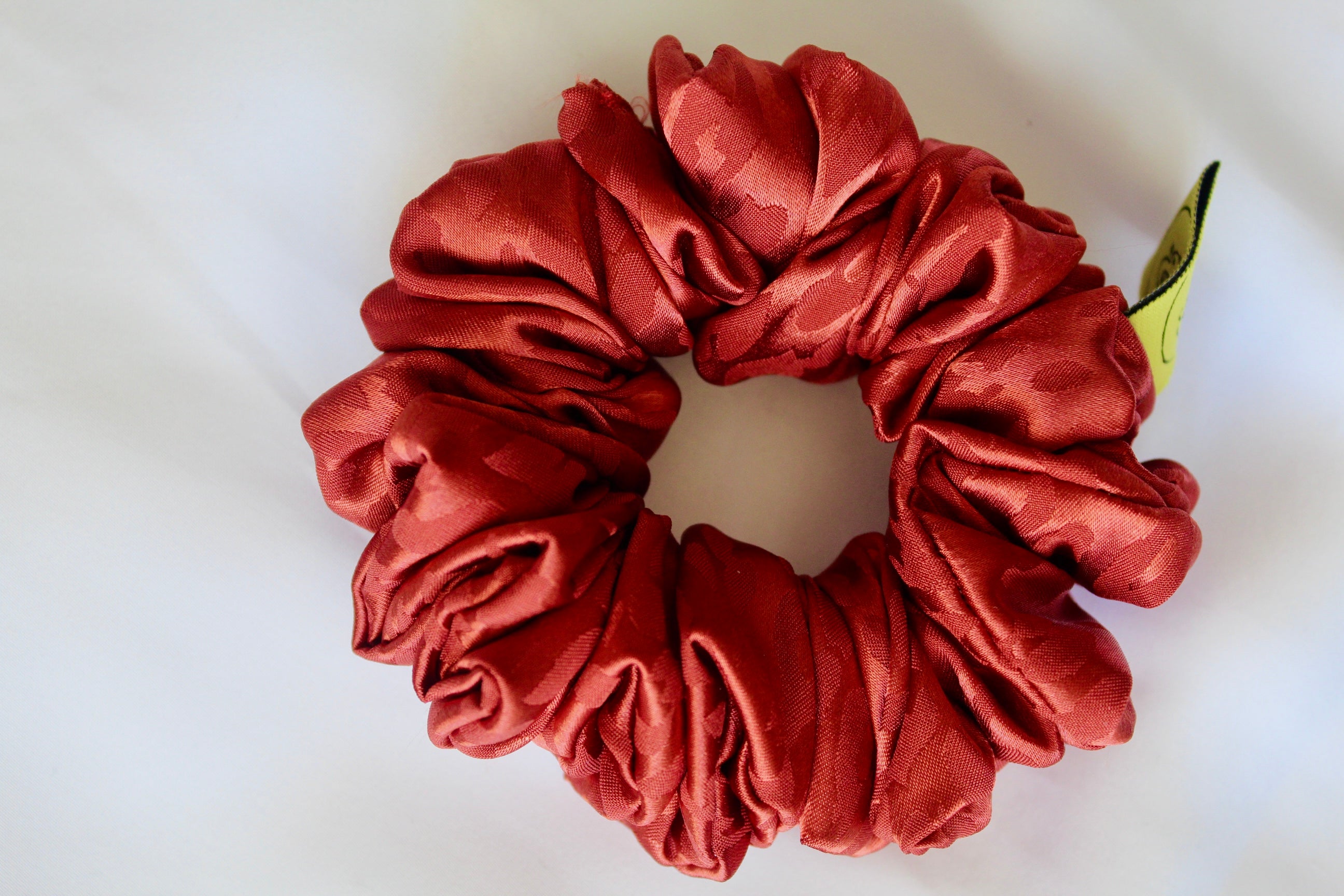 This shows a luxurious scrunchie made of silky textured fabric in a burnt orange/red colour.The fabric has a subtle leopard print pattern wove into it, giving it an elegant and slightly reflective appearance.The scrunchie is gathered and voluminous creating soft folds and a plush look.A small yellow label tag with a picture of a duck is attached to the scrunchie adding a touch of branding.The overall aesthetic is soft, elegant and stylish. Perfect for those with sensitive scalp or hair loss