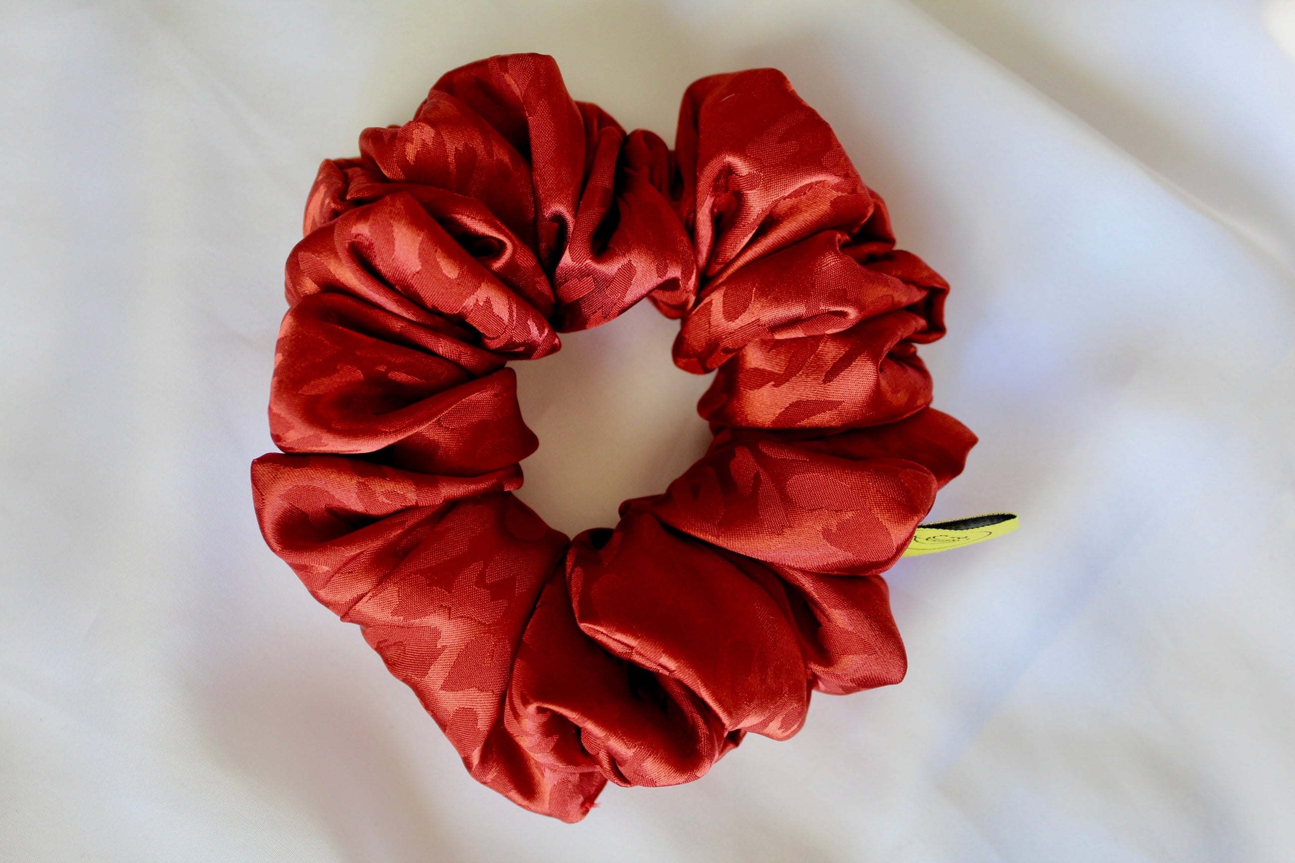 This shows a luxurious scrunchie made of silky textured fabric in a burnt orange/red colour.The fabric has a subtle leopard print pattern wove into it, giving it an elegant and slightly reflective appearance.The scrunchie is gathered and voluminous creating soft folds and a plush look.A small yellow label tag with a picture of a duck is attached to the scrunchie adding a touch of branding.The overall aesthetic is soft, elegant and stylish. Perfect for those with sensitive scalp or hair loss