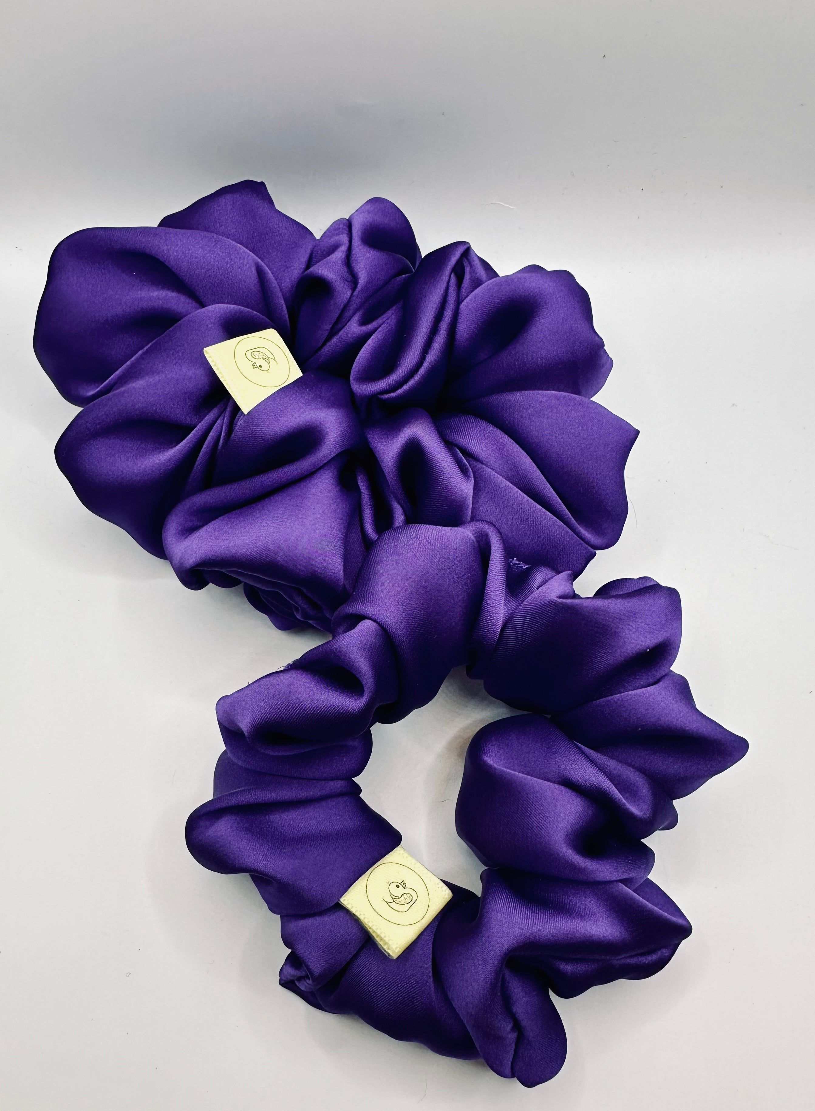 This shows a luxurious scrunchie made of silky textured fabric in a deep purple colour giving it an elegant and slightly reflective appearance.The scrunchie is gathered and voluminous creating soft folds and a plush look.A small yellow label tag with a picture of a duck is attached to the scrunchie adding a touch of branding.The overall aesthetic is soft, elegant and stylish. Perfect for those with sensitive scalp or hair loss