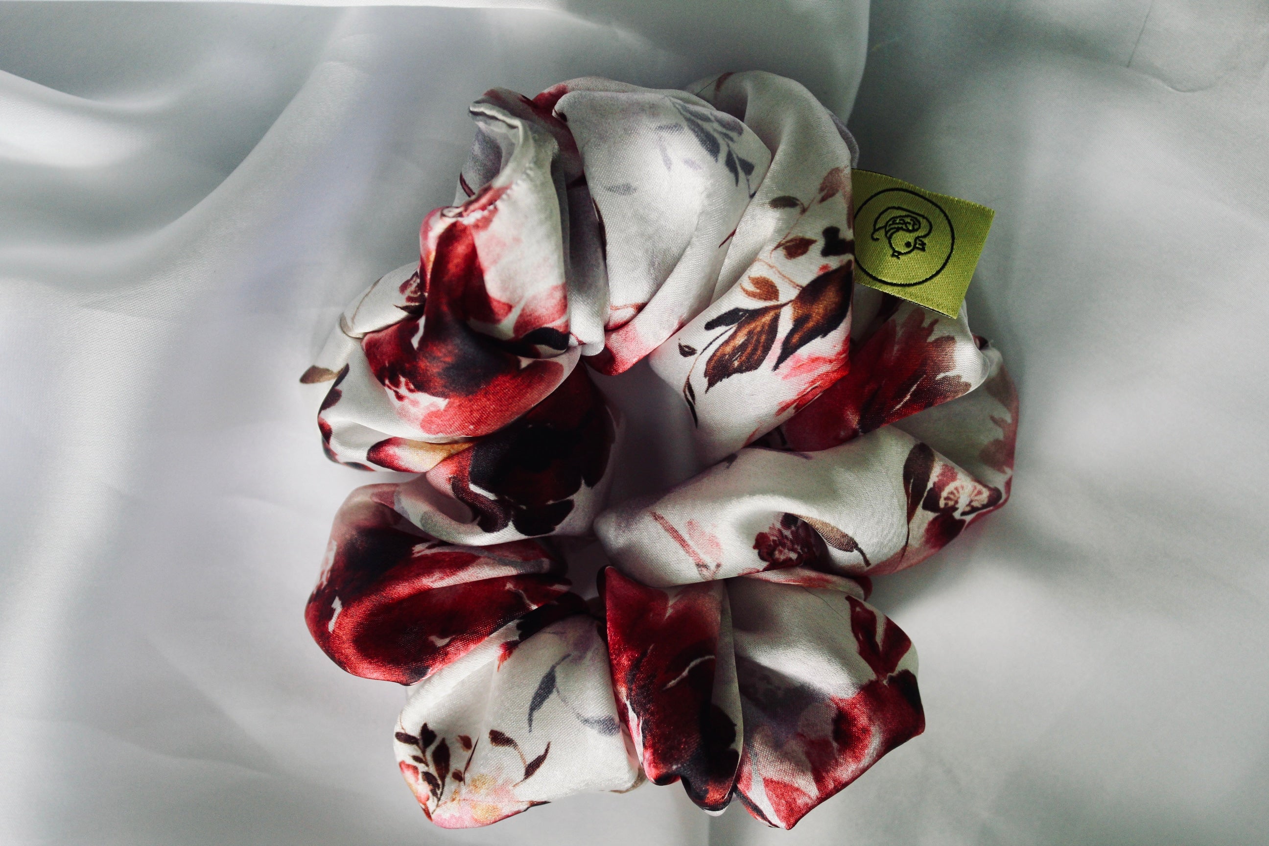 This shows a luxurious scrunchie made of silky textured fabric in a dusty pink and red floral pattern. The fabric has a subtle floral pattern wove into it, giving it an elegant and slightly reflective appearance.The scrunchie is gathered and voluminous creating soft folds and a plush look.A small yellow label tag with a picture of a duck is attached to the scrunchie adding a touch of branding.The overall aesthetic is soft, elegant and stylish. Perfect for those with sensitive scalp or hair loss