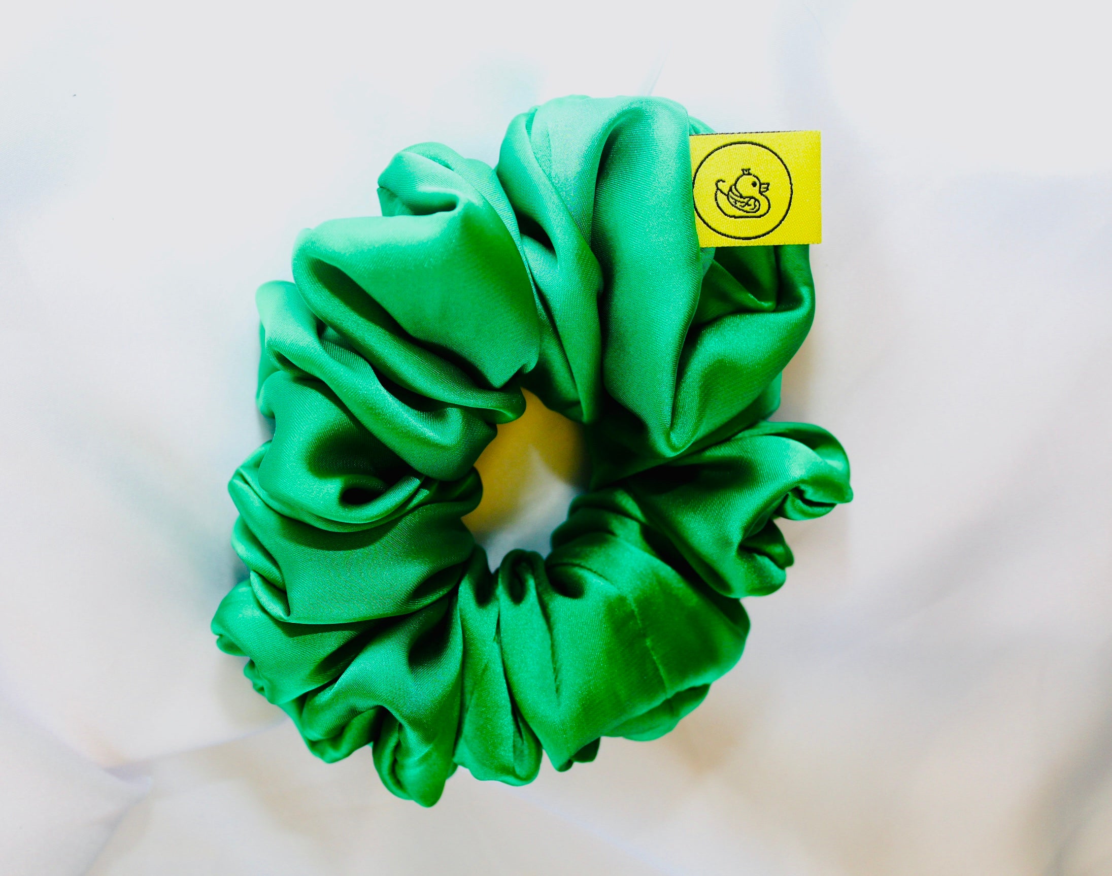 This shows a luxurious scrunchie made of silky textured fabric in an Emerald Green giving it an elegant and slightly reflective appearance.The scrunchie is gathered and voluminous creating soft folds and a plush look.A small yellow label tag with a picture of a duck is attached to the scrunchie adding a touch of branding.The overall aesthetic is soft, elegant and stylish. Perfect for those with sensitive scalp or hair loss
