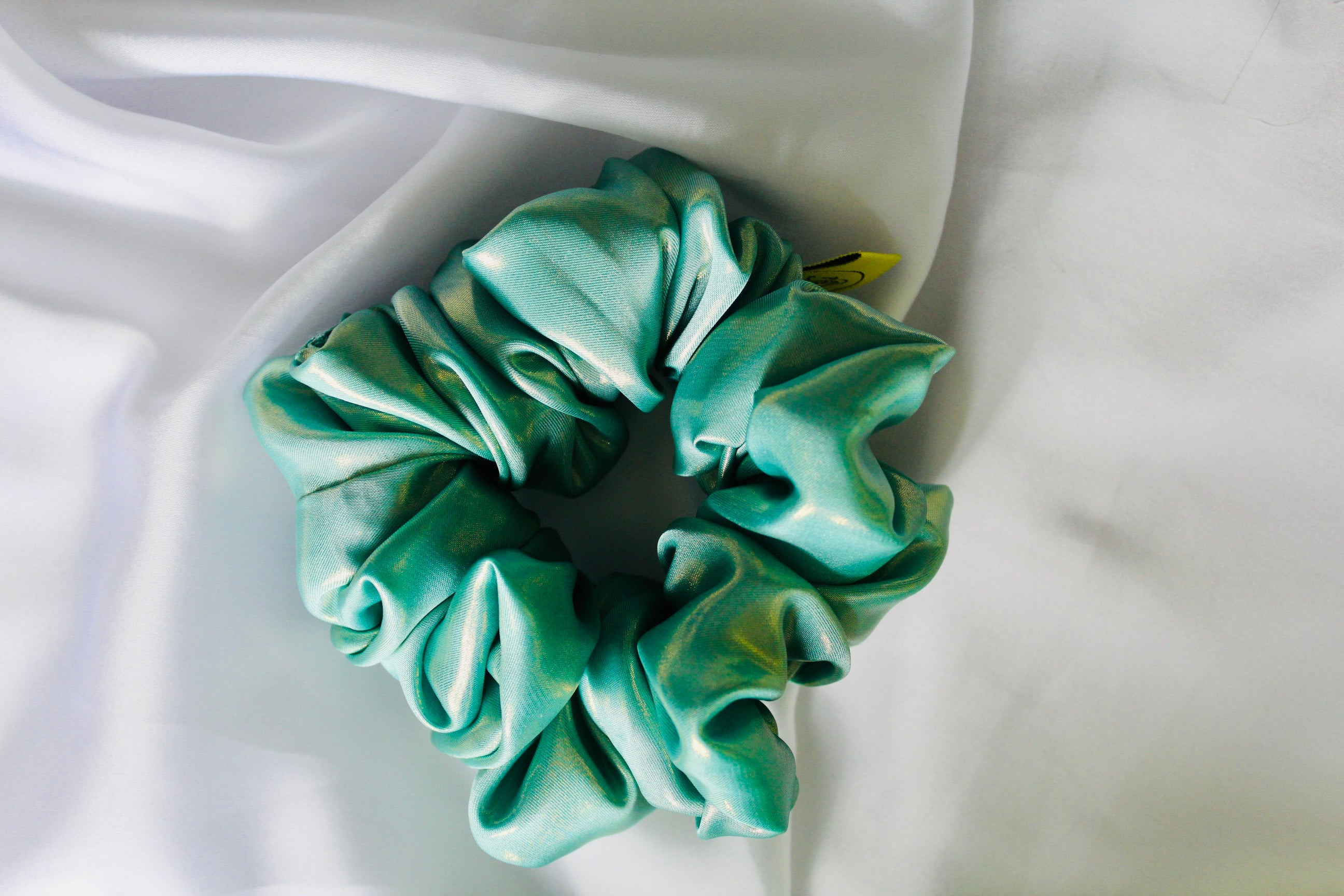 This shows a luxurious scrunchie made of silky textured fabric in an Aqua Green with a liquid gold metallic running through the fabric giving it an elegant and slightly reflective appearance.The scrunchie is gathered and voluminous creating soft folds and a plush look.A small yellow label tag with a picture of a duck is attached to the scrunchie adding a touch of branding.The overall aesthetic is soft, elegant and stylish. Perfect for those with sensitive scalp or hair loss