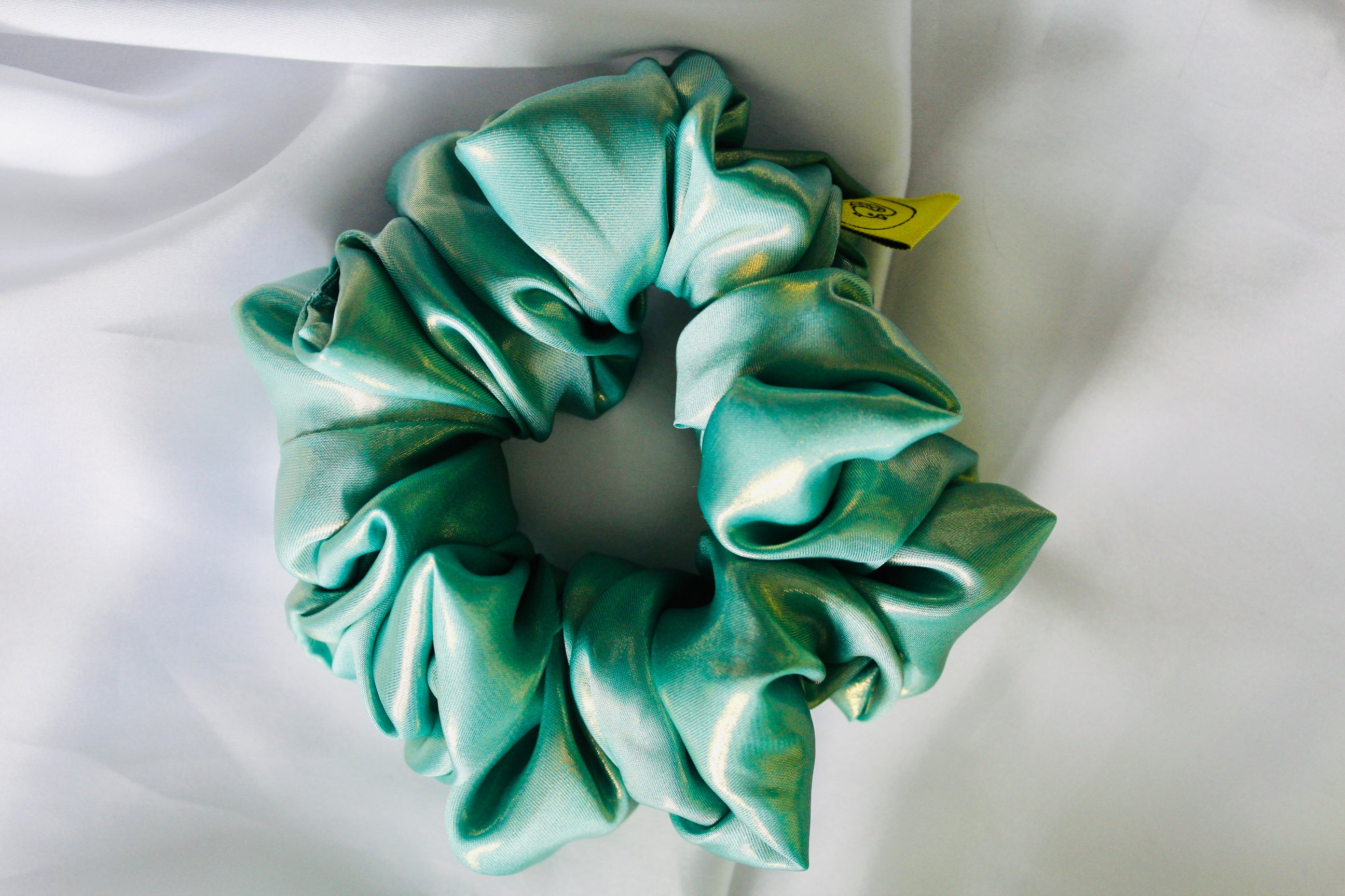 This shows a luxurious scrunchie made of silky textured fabric in an Aqua Metallic Gold The fabric has a subtle floral pattern, giving it an elegant and slightly reflective appearance.The scrunchie is gathered and voluminous creating soft folds and a plush look.A small yellow label tag with a picture of a duck is attached to the scrunchie adding a touch of branding.The overall aesthetic is soft, elegant and stylish. Perfect for those with sensitive scalp or hair loss
