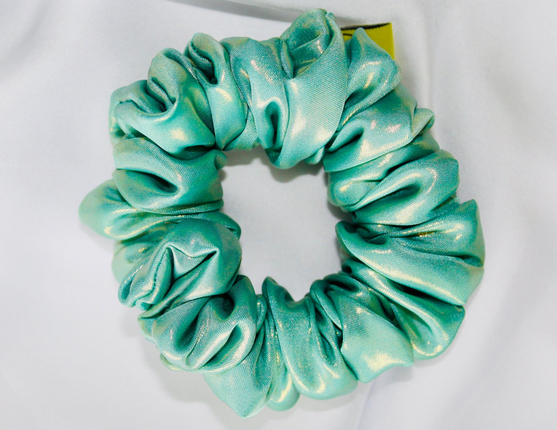 This shows a luxurious scrunchie made of silky textured fabric in an Aqua Metallic Gold The fabric has a subtle floral pattern, giving it an elegant and slightly reflective appearance.The scrunchie is gathered and voluminous creating soft folds and a plush look.A small yellow label tag with a picture of a duck is attached to the scrunchie adding a touch of branding.The overall aesthetic is soft, elegant and stylish. Perfect for those with sensitive scalp or hair loss