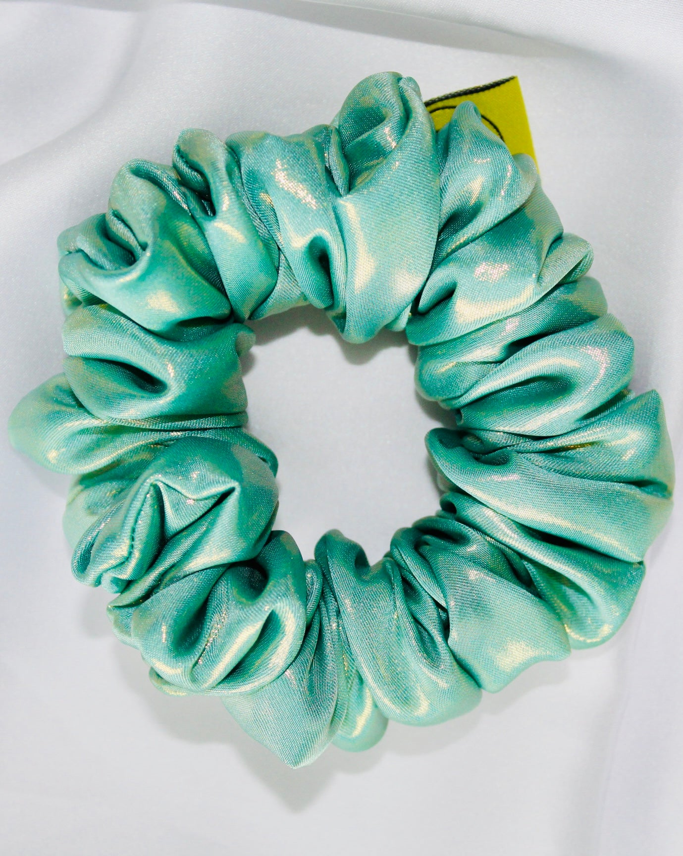 This shows a luxurious scrunchie made of silky textured fabric in an Aqua Metallic Gold The fabric has a subtle floral pattern, giving it an elegant and slightly reflective appearance.The scrunchie is gathered and voluminous creating soft folds and a plush look.A small yellow label tag with a picture of a duck is attached to the scrunchie adding a touch of branding.The overall aesthetic is soft, elegant and stylish. Perfect for those with sensitive scalp or hair loss