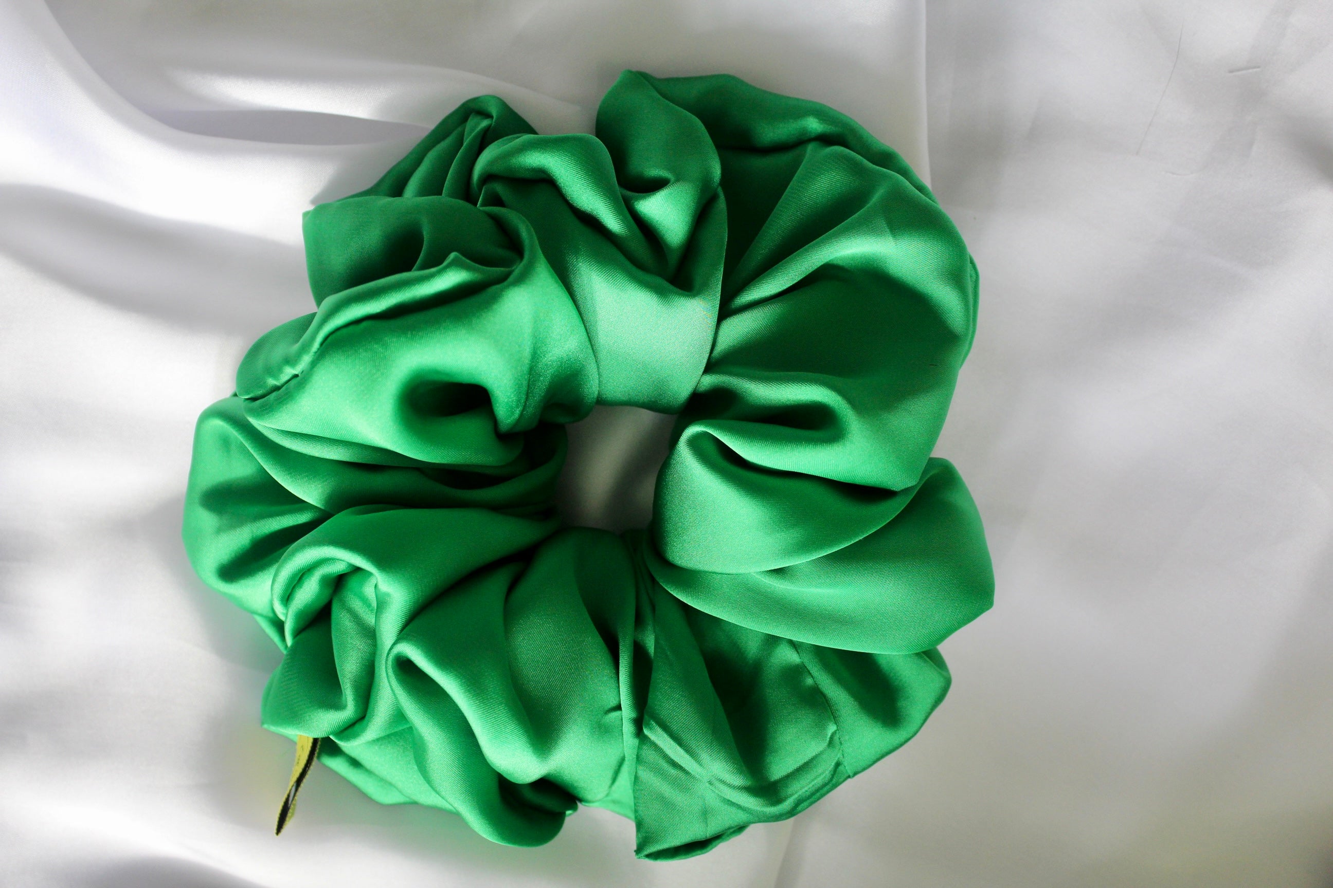 This shows a luxurious scrunchie made of silky textured fabric in an Emerald Green giving it an elegant and slightly reflective appearance.The scrunchie is gathered and voluminous creating soft folds and a plush look.A small yellow label tag with a picture of a duck is attached to the scrunchie adding a touch of branding.The overall aesthetic is soft, elegant and stylish. Perfect for those with sensitive scalp or hair loss