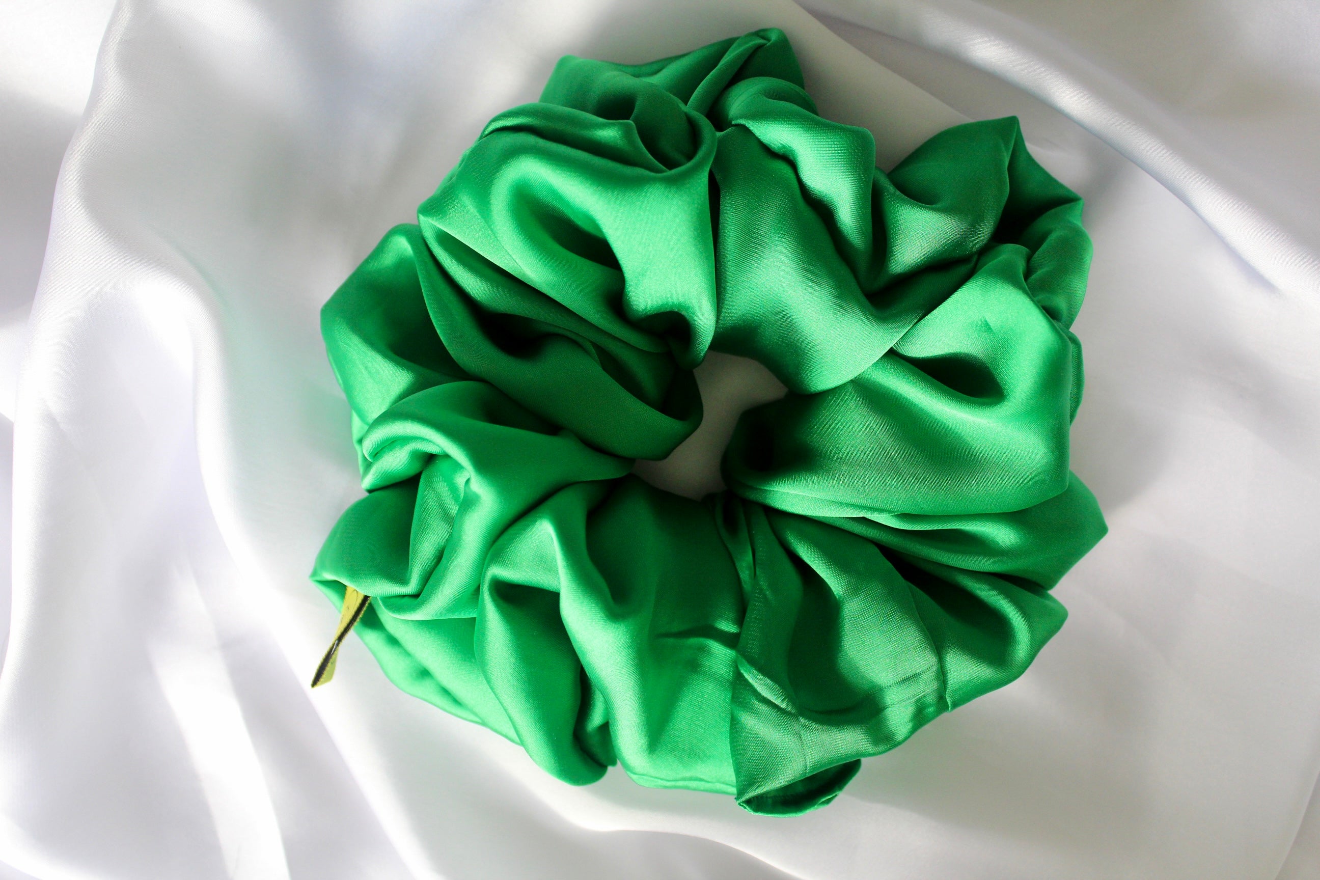 This shows a luxurious scrunchie made of silky textured fabric in a Emerald Green giving it an elegant and slightly reflective appearance.The scrunchie is gathered and voluminous creating soft folds and a plush look.A small yellow label tag with a picture of a duck is attached to the scrunchie adding a touch of branding.The overall aesthetic is soft, elegant and stylish. Perfect for those with sensitive scalp or hair loss