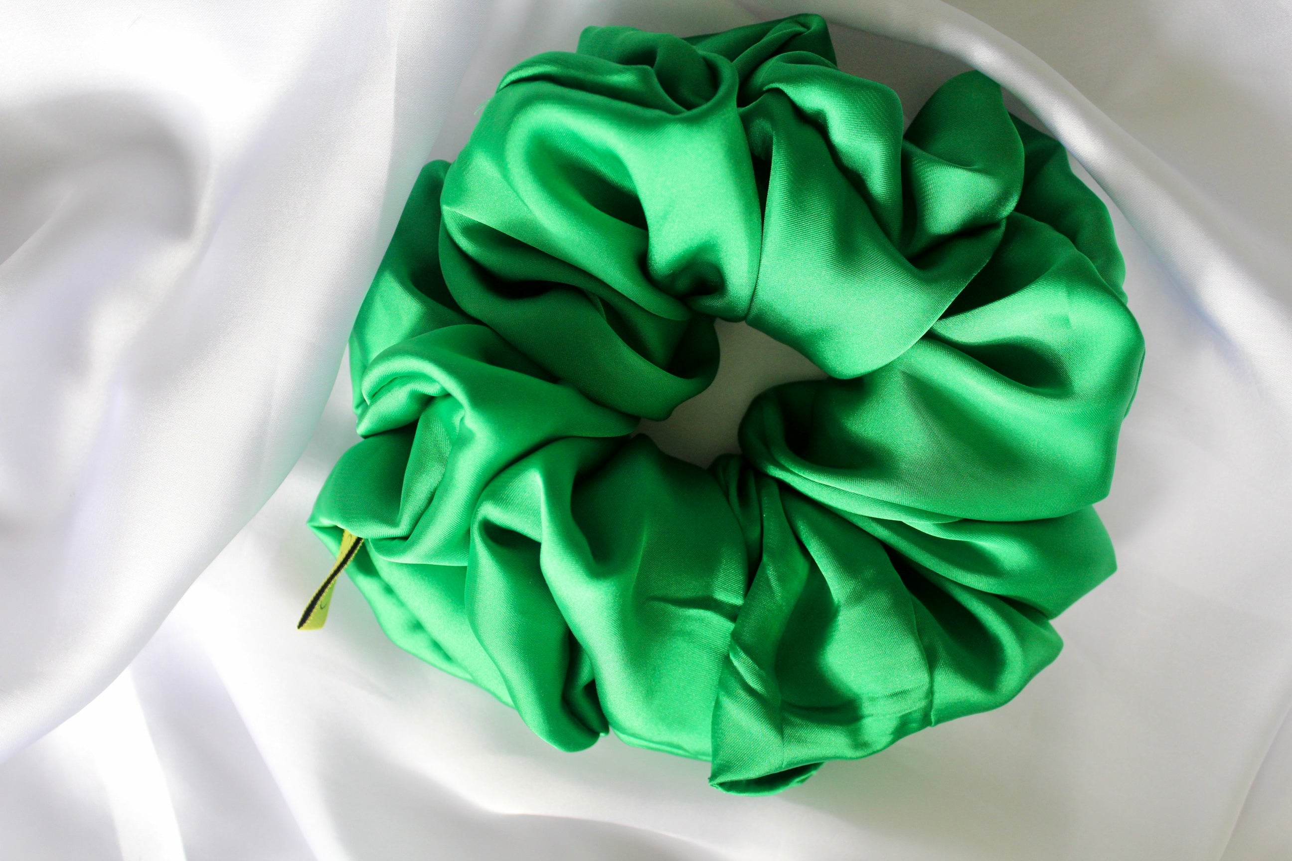 This shows a luxurious scrunchie made of silky textured fabric in a Emerald Green giving it an elegant and slightly reflective appearance.The scrunchie is gathered and voluminous creating soft folds and a plush look.A small yellow label tag with a picture of a duck is attached to the scrunchie adding a touch of branding.The overall aesthetic is soft, elegant and stylish. Perfect for those with sensitive scalp or hair loss