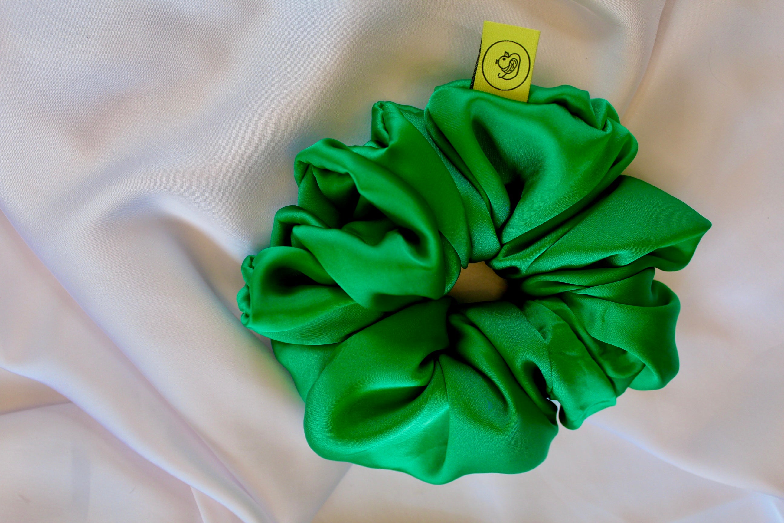 This shows a luxurious scrunchie made of silky textured fabric in a Emerald Green giving it an elegant and slightly reflective appearance.The scrunchie is gathered and voluminous creating soft folds and a plush look.A small yellow label tag with a picture of a duck is attached to the scrunchie adding a touch of branding.The overall aesthetic is soft, elegant and stylish. Perfect for those with sensitive scalp or hair loss