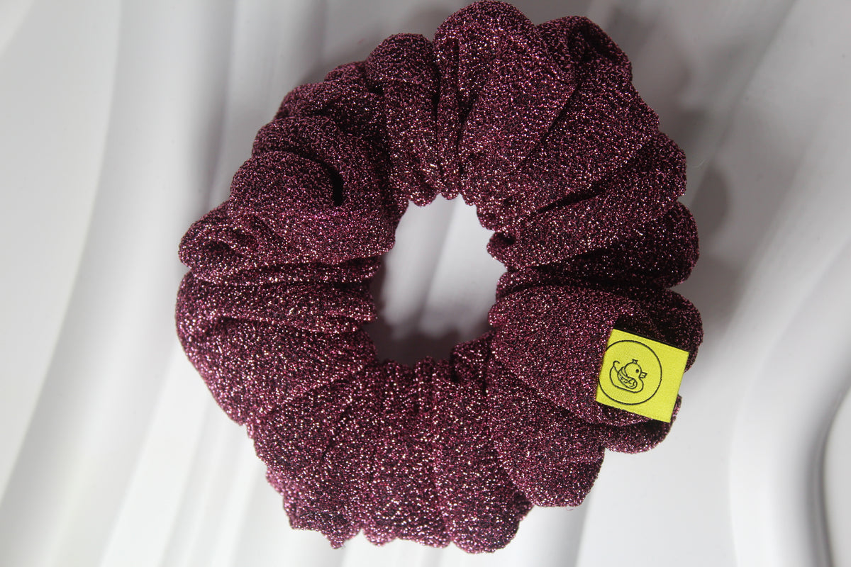 A glittery burgundy scrunchie with a small yellow brand tag, photographed on a draped white fabric background.