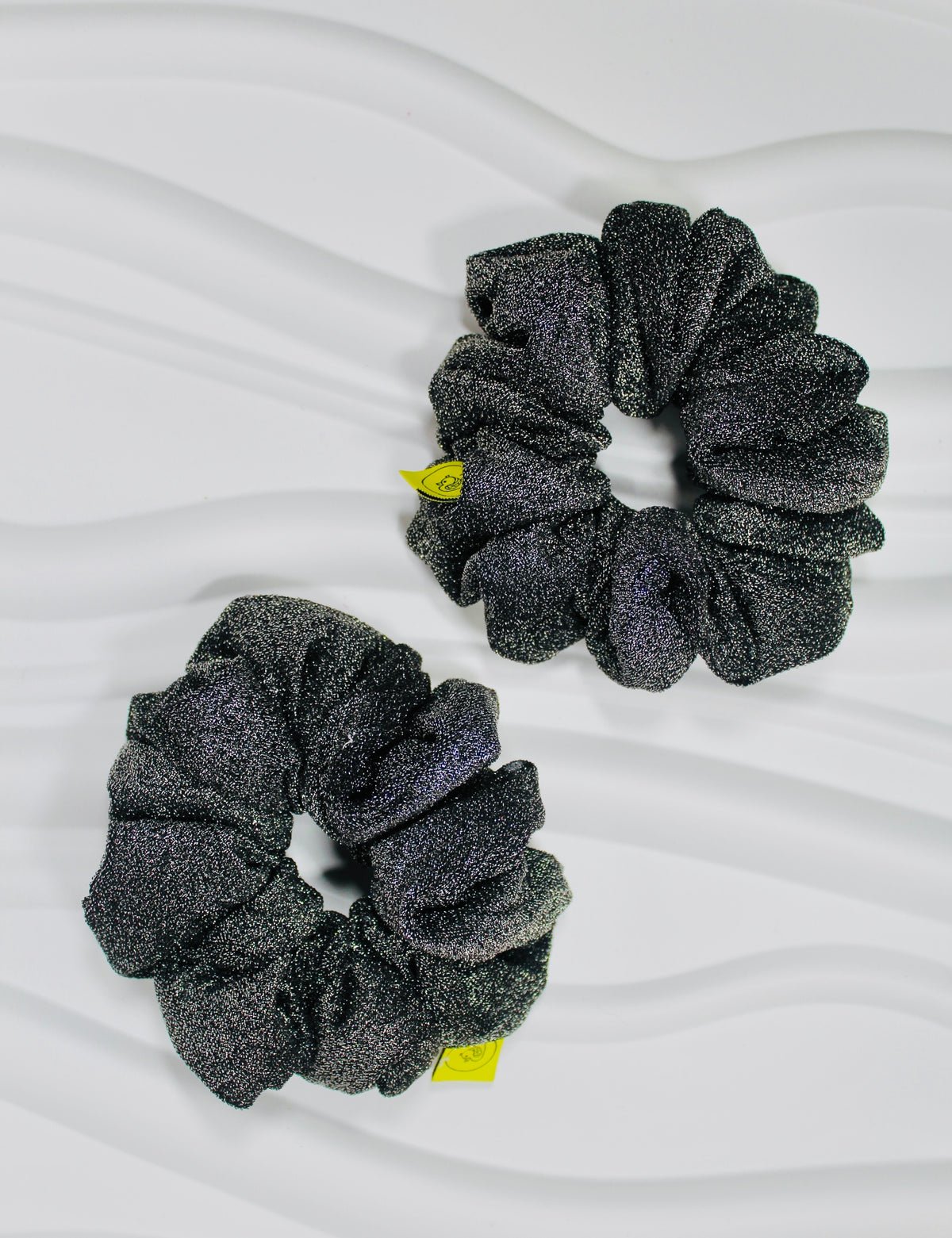 Two dark gray glittery scrunchies with small yellow tags, displayed on a white wavy background.