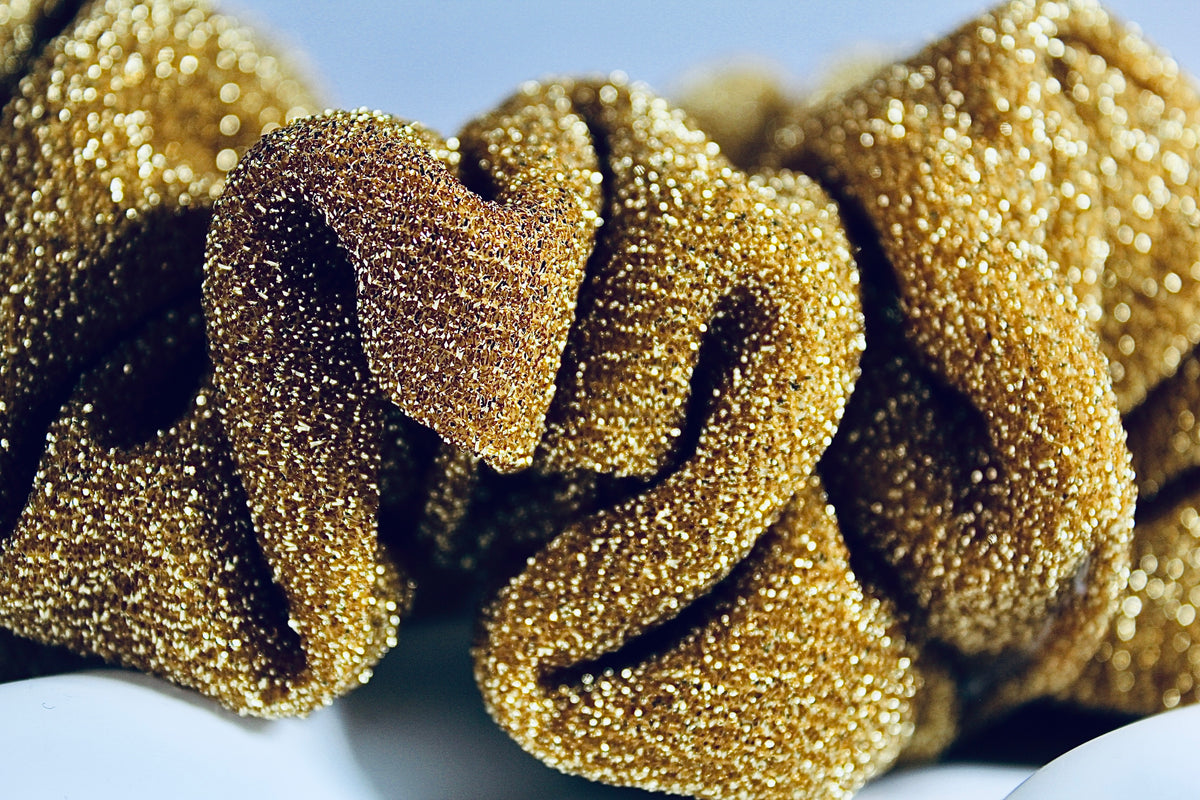 A close-up of a gold glitter scrunchie with a textured, sparkly surface, resting on a white surface against a light blue background.