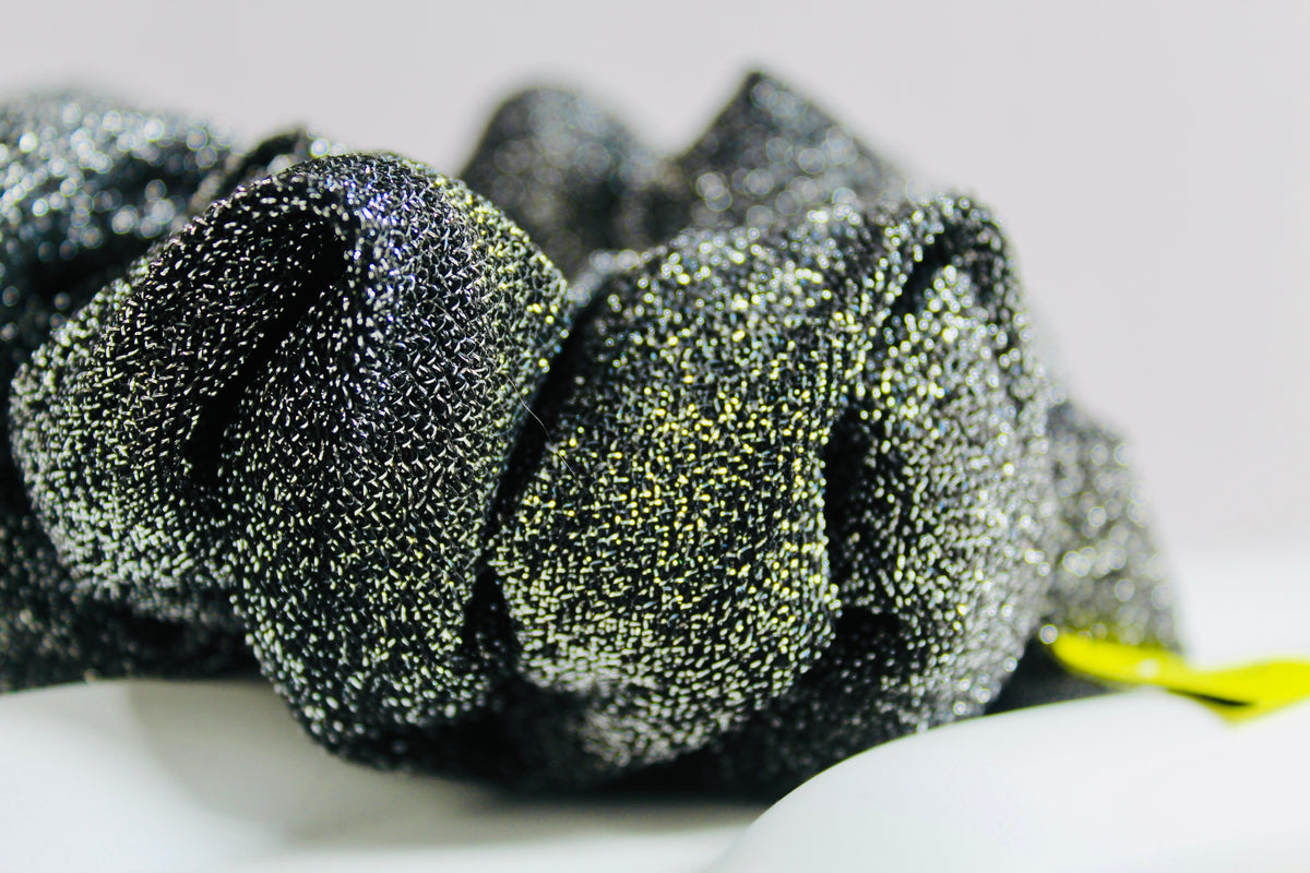 A close-up of a black glittery fabric scrunchie with fine metallic flecks, resting on a white surface against a light gray background.