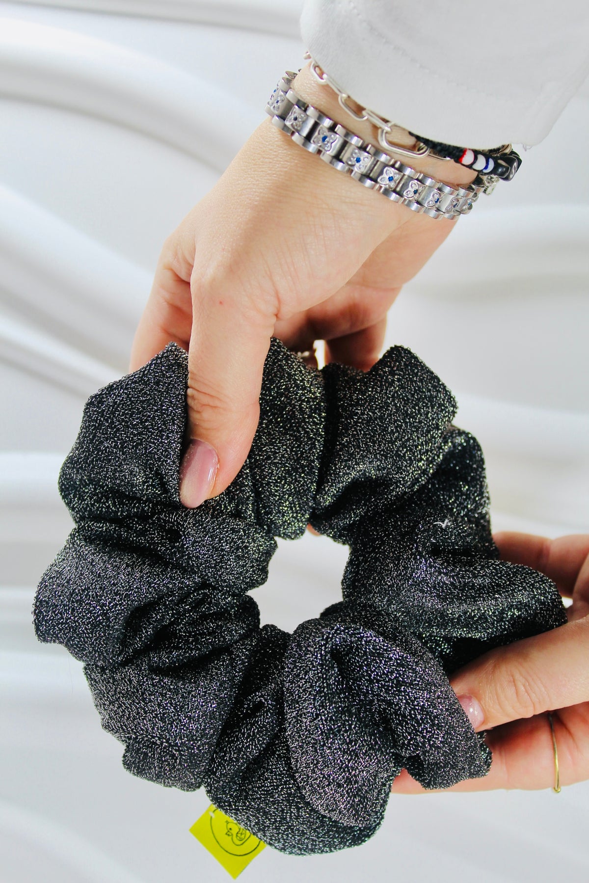 A close-up of a person holding a dark glittery scrunchie with both hands against a soft white fabric background. The scrunchie has a sparkling charcoal gray to black finish with subtle purple undertones.