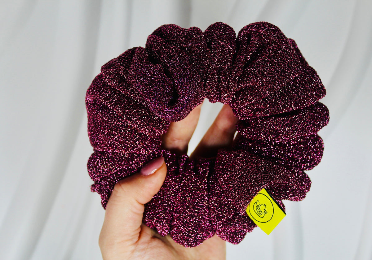 A close-up of a hand holding a dark purple glittery scrunchie against a soft white fabric background. A small yellow tag with a smiley face is attached to the scrunchie.