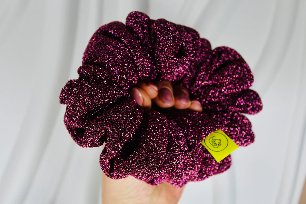 A close-up of a hand holding a large, glittery magenta scrunchie against a soft white draped fabric background. A small yellow tag is attached to the scrunchie.