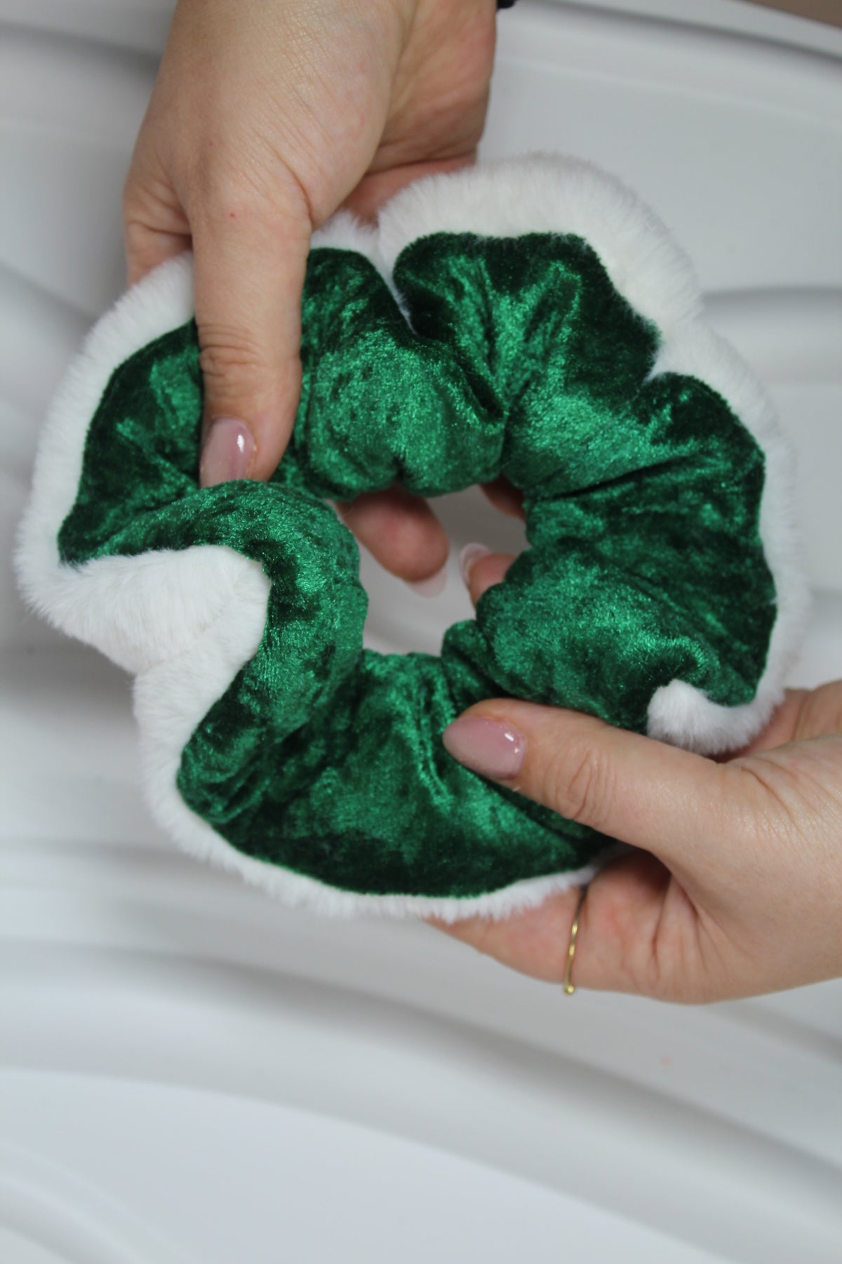 A green velvet scrunchie with a white faux fur trim, held by two hands against a white background.