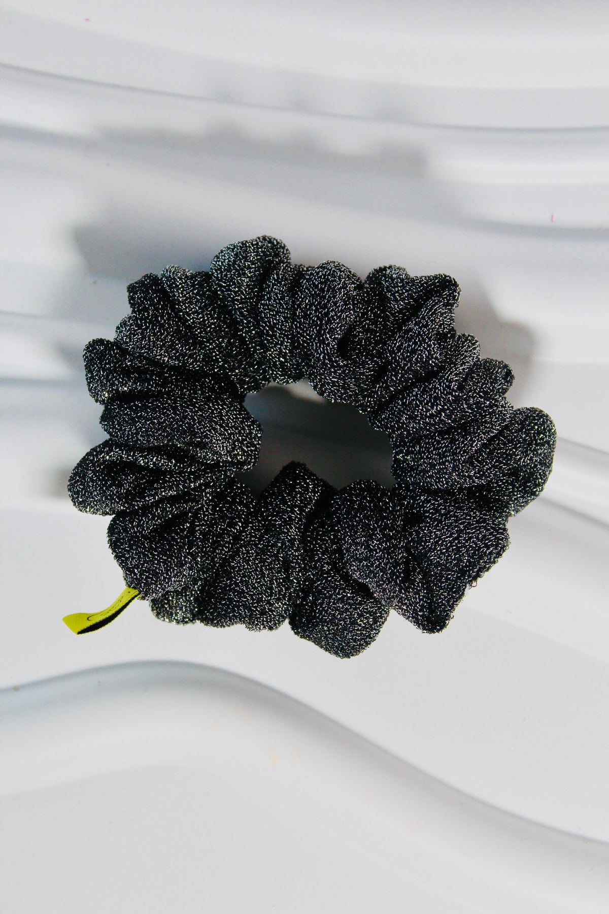 A black glittery scrunchie with a small lime green ribbon tail, photographed on a wavy white surface.