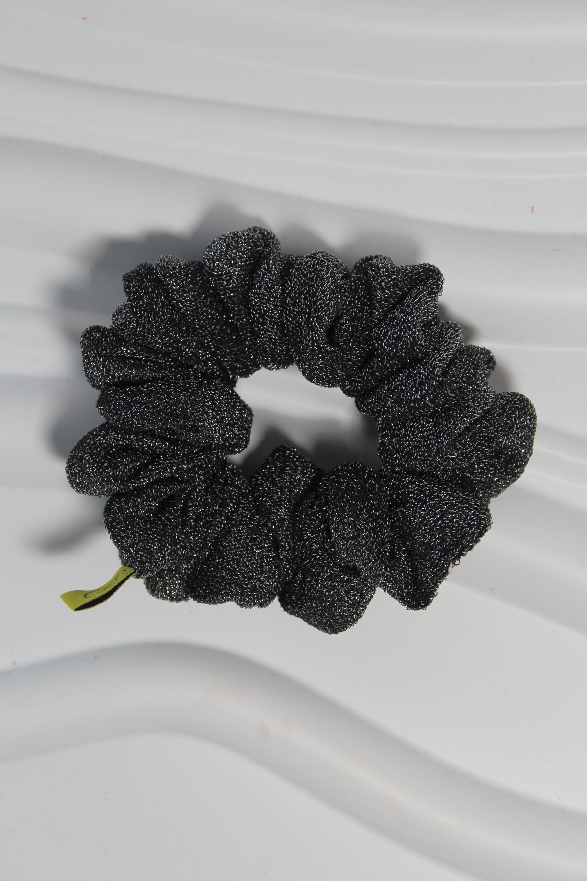 A black glittery scrunchie with a small green tag, displayed on a white wavy surface.