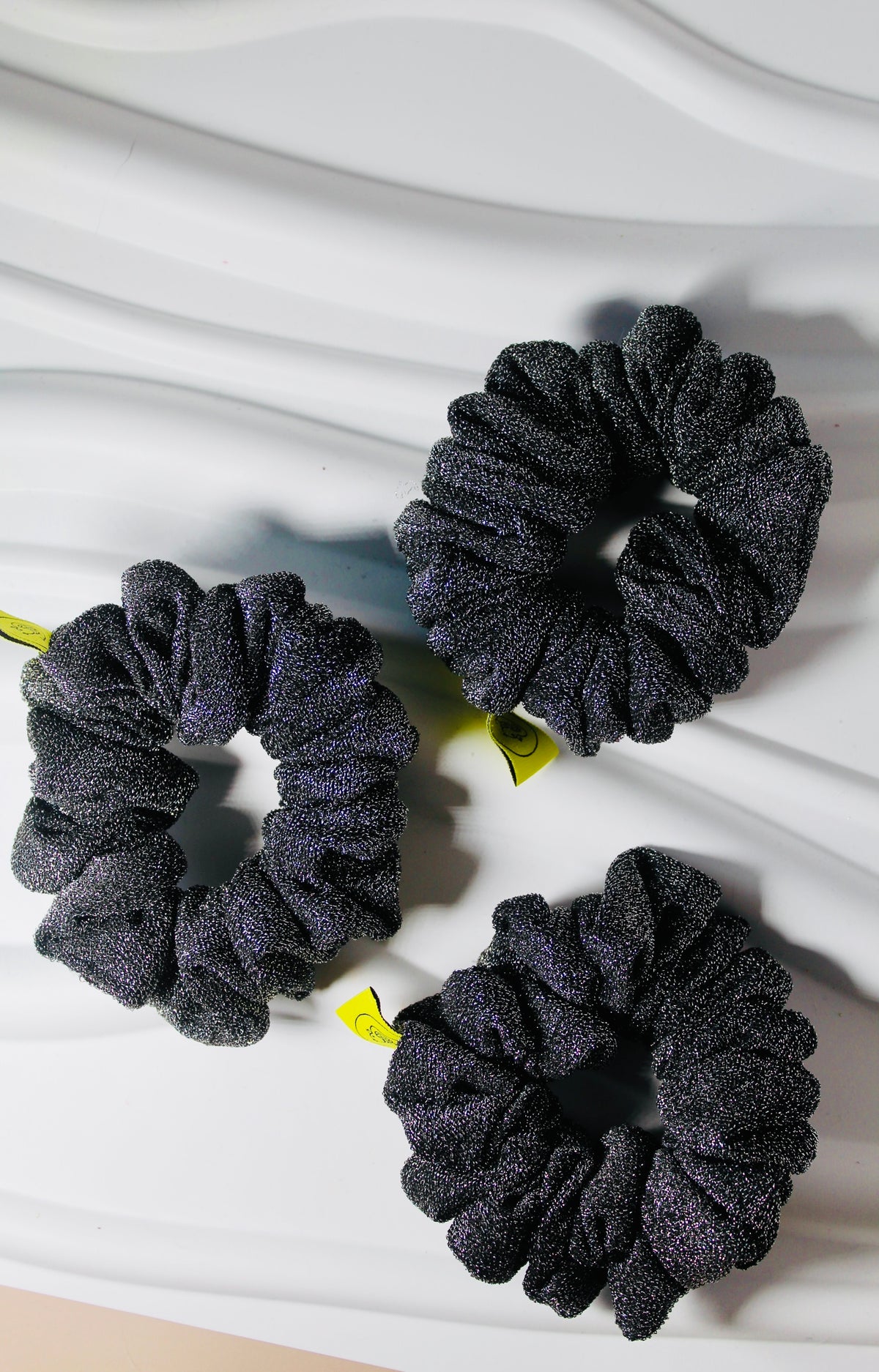 Three dark gray glittery scrunchies with small yellow ribbon tags, arranged on a white wavy background.
