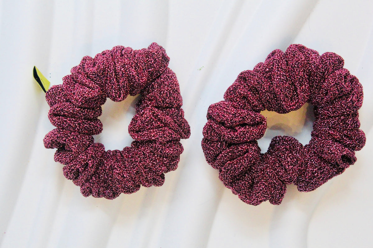 Two textured, glittery deep magenta scrunchies on a white draped fabric background.