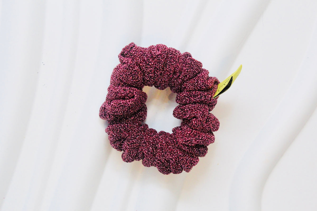 A textured, sparkly deep burgundy scrunchie with a small yellow tag, displayed on a white wavy background.