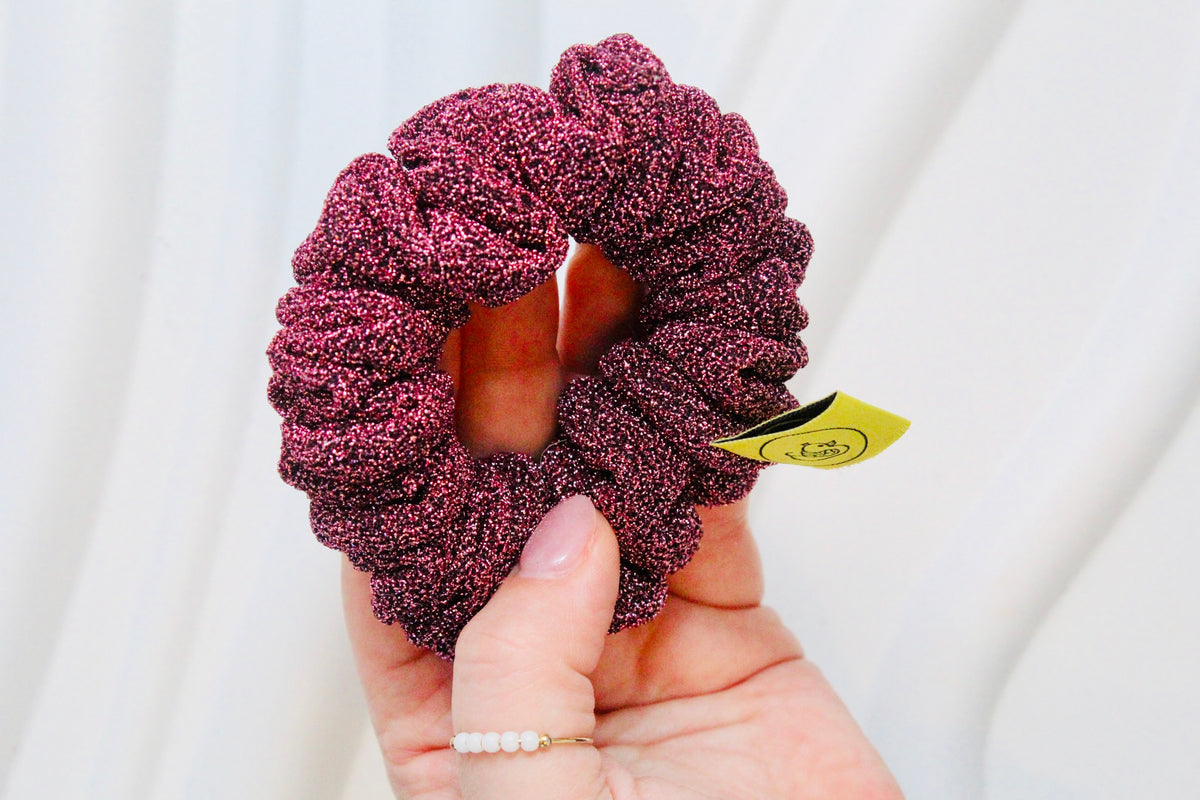 A close-up of a hand holding a textured, sparkly deep burgundy scrunchie with a small yellow tag attached, set against a soft, draped white fabric background.