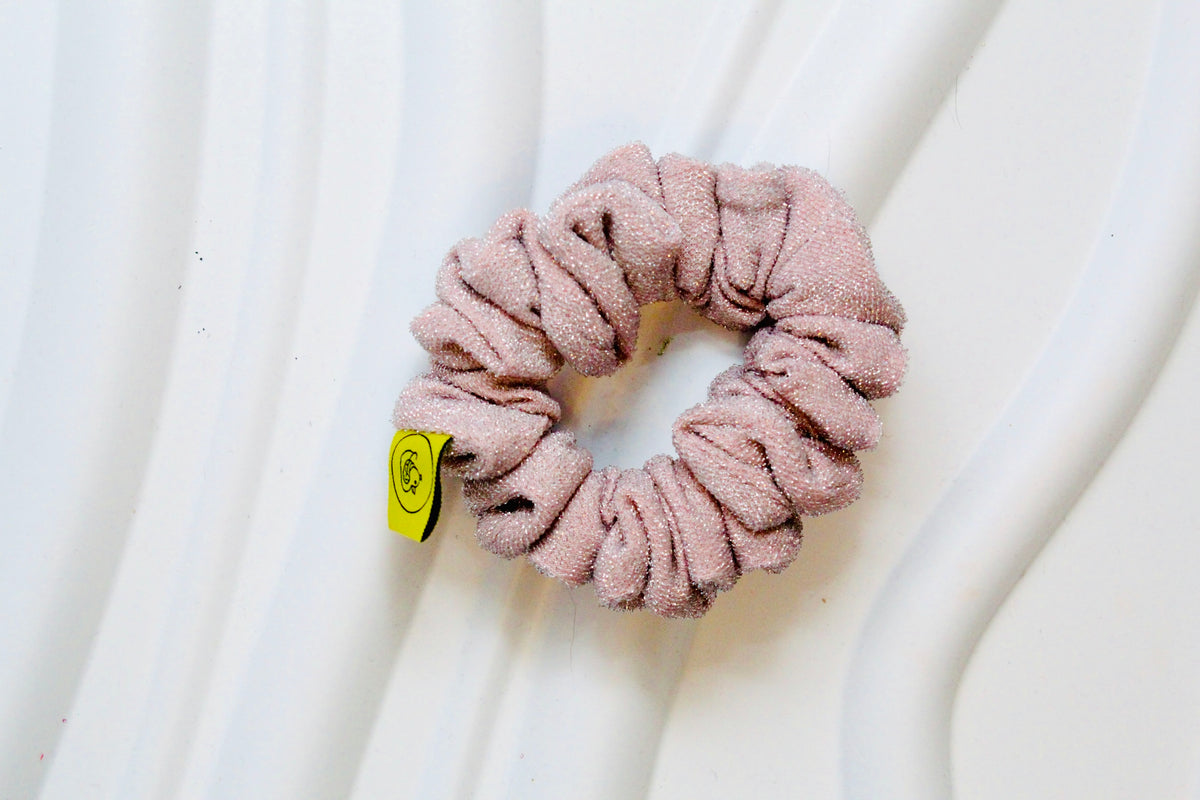 A dusty pink scrunchie made from textured terry cloth, featuring a small yellow brand tag on the side, displayed on a white wavy surface.