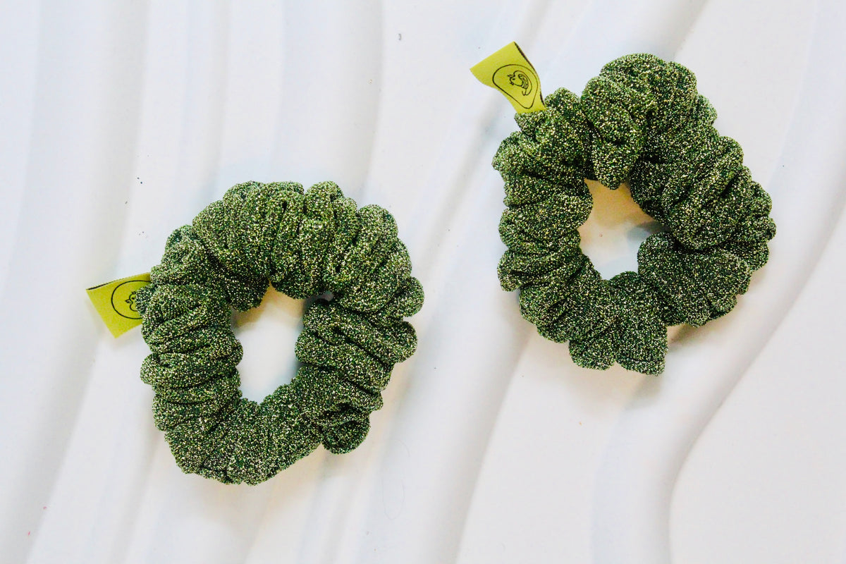 Two green glittery scrunchies with small yellow tags, displayed on a white wavy surface.