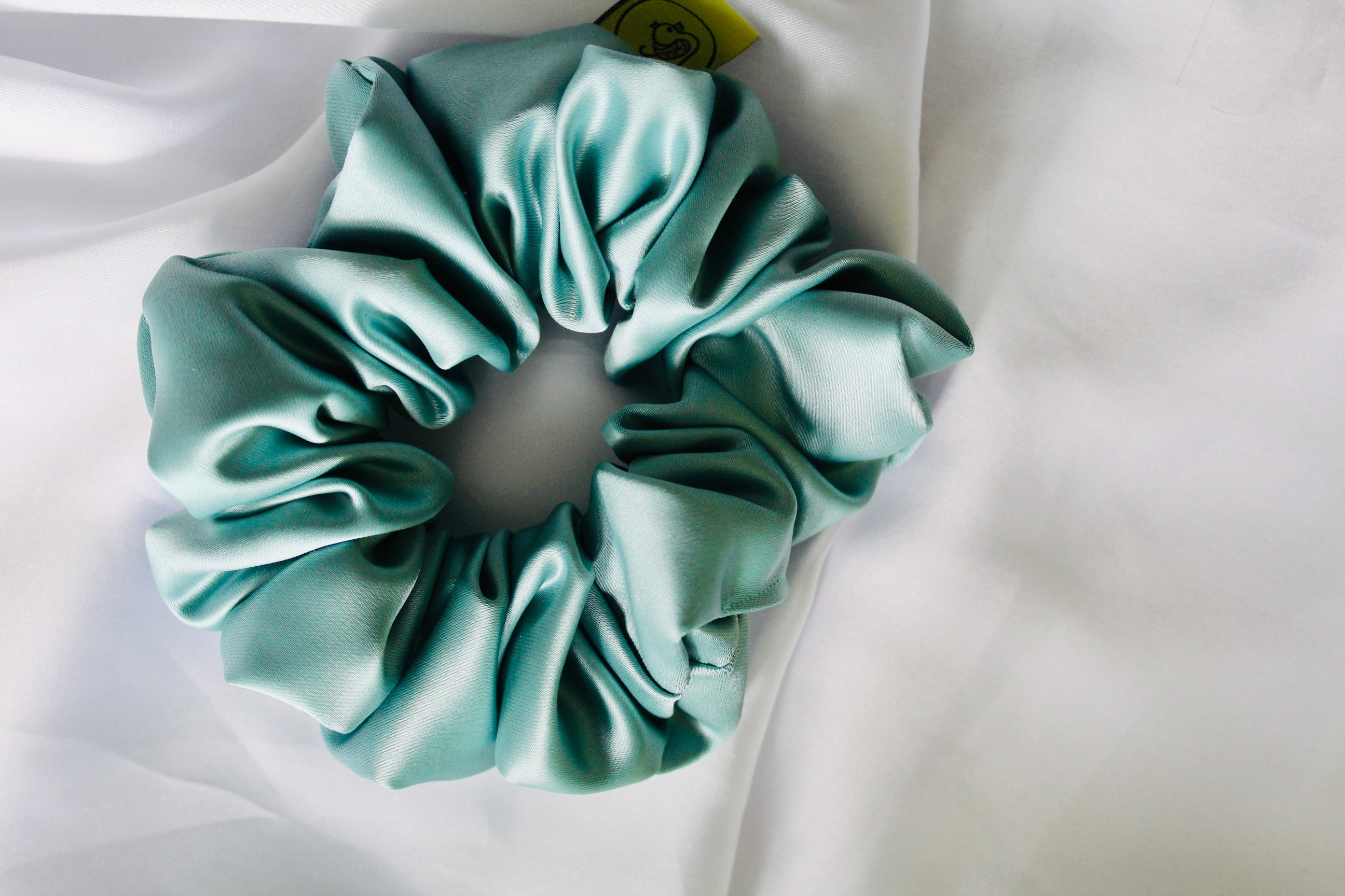 This shows a luxurious scrunchie made of silky textured fabric in an mint green running through the fabric giving it an elegant and slightly reflective appearance.The scrunchie is gathered and voluminous creating soft folds and a plush look.A small yellow label tag with a picture of a duck is attached to the scrunchie adding a touch of branding.The overall aesthetic is soft, elegant and stylish. Perfect for those with sensitive scalp or hair loss
