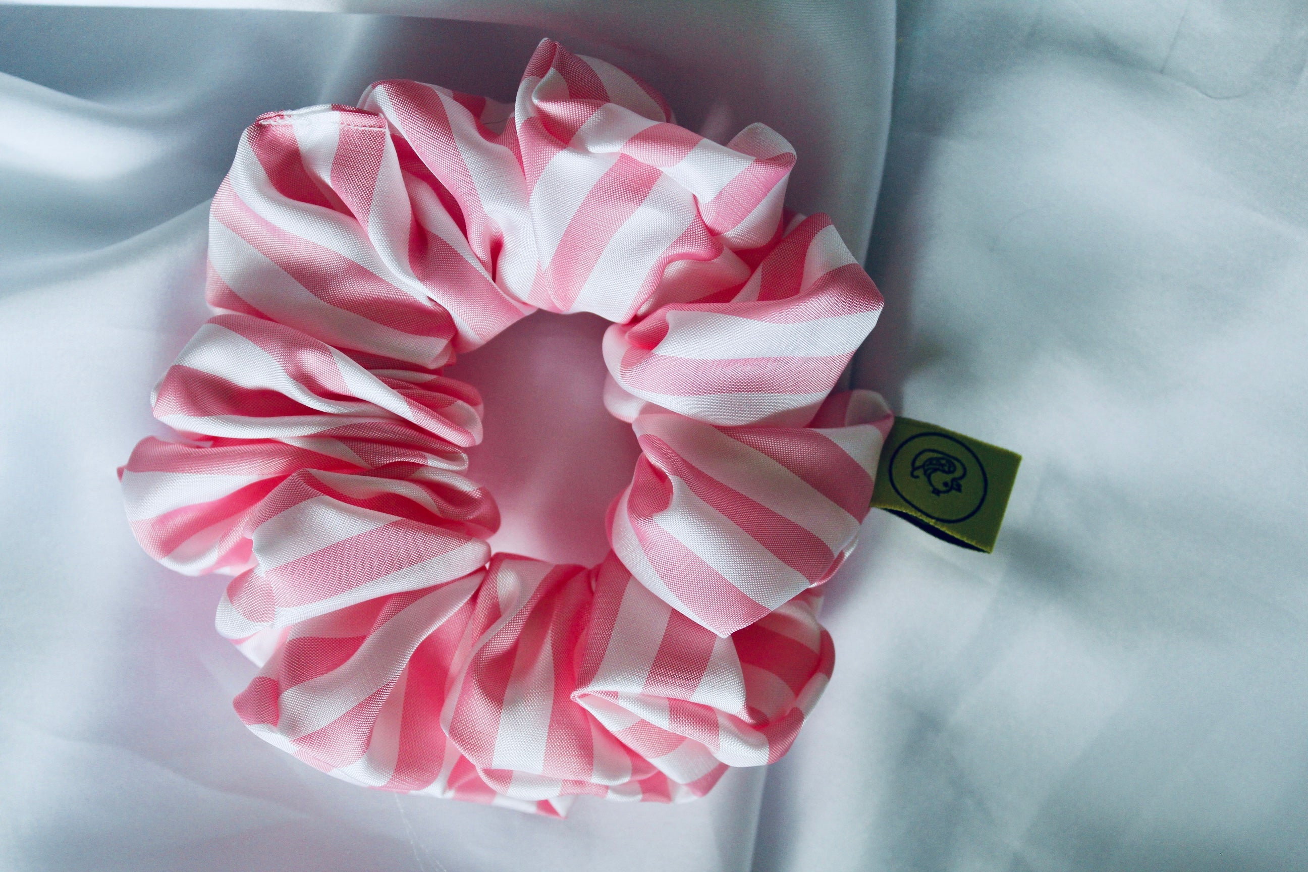 This shows a luxurious scrunchie made of silky textured fabric in a Baby pink and white striped colour, giving it an elegant and slightly reflective appearance.The scrunchie is gathered and voluminous creating soft folds and a plush look.A small yellow label tag with a picture of a duck is attached to the scrunchie adding a touch of branding.The overall aesthetic is soft, elegant and stylish. Perfect for those with sensitive scalp or hair loss