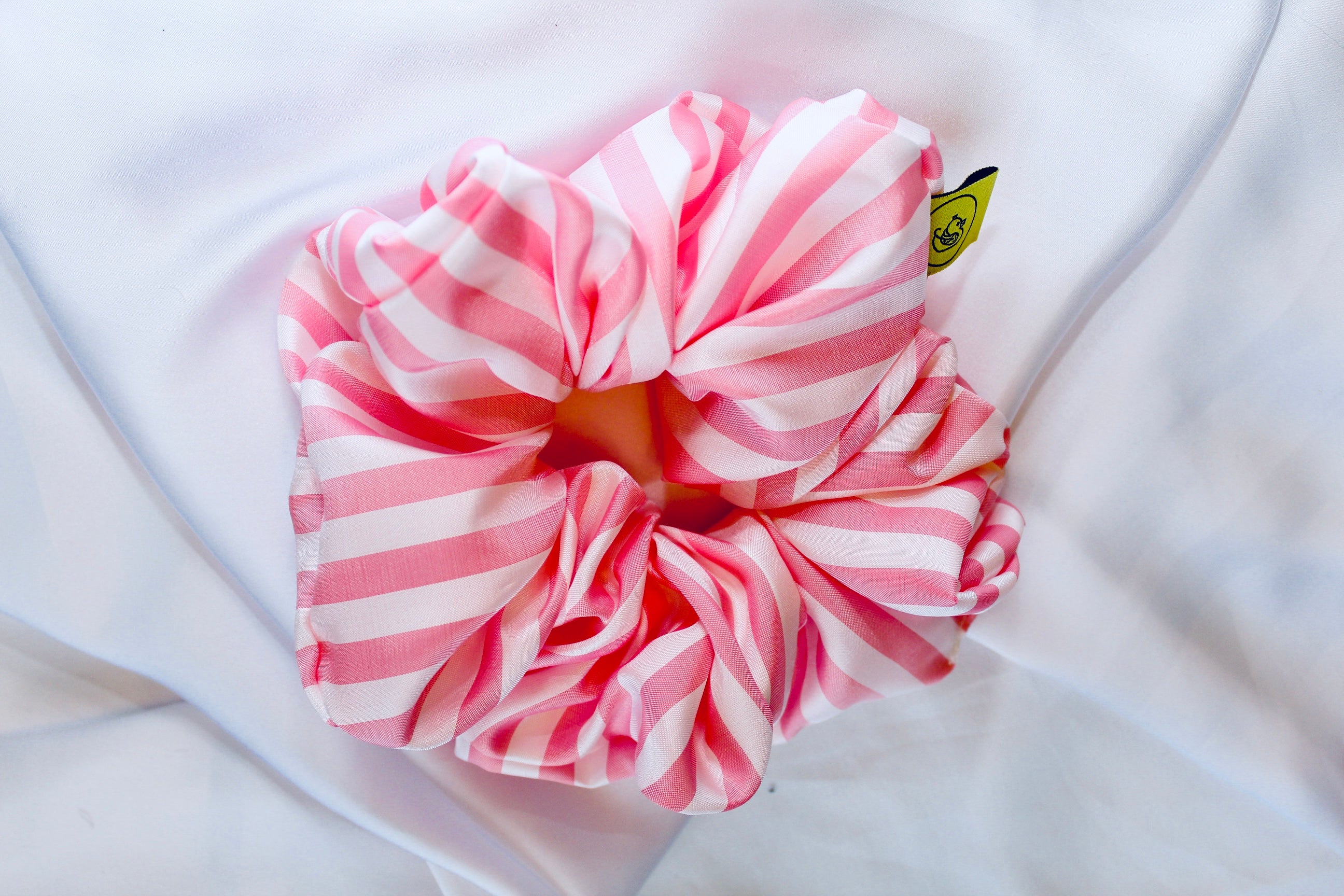 This shows a luxurious scrunchie made of silky textured fabric in a baby pink and white stripe pattern through the fabric giving it an elegant and slightly reflective appearance.The scrunchie is gathered and voluminous creating soft folds and a plush look.A small yellow label tag with a picture of a duck is attached to the scrunchie adding a touch of branding.The overall aesthetic is soft, elegant and stylish. Perfect for those with sensitive scalp or hair loss