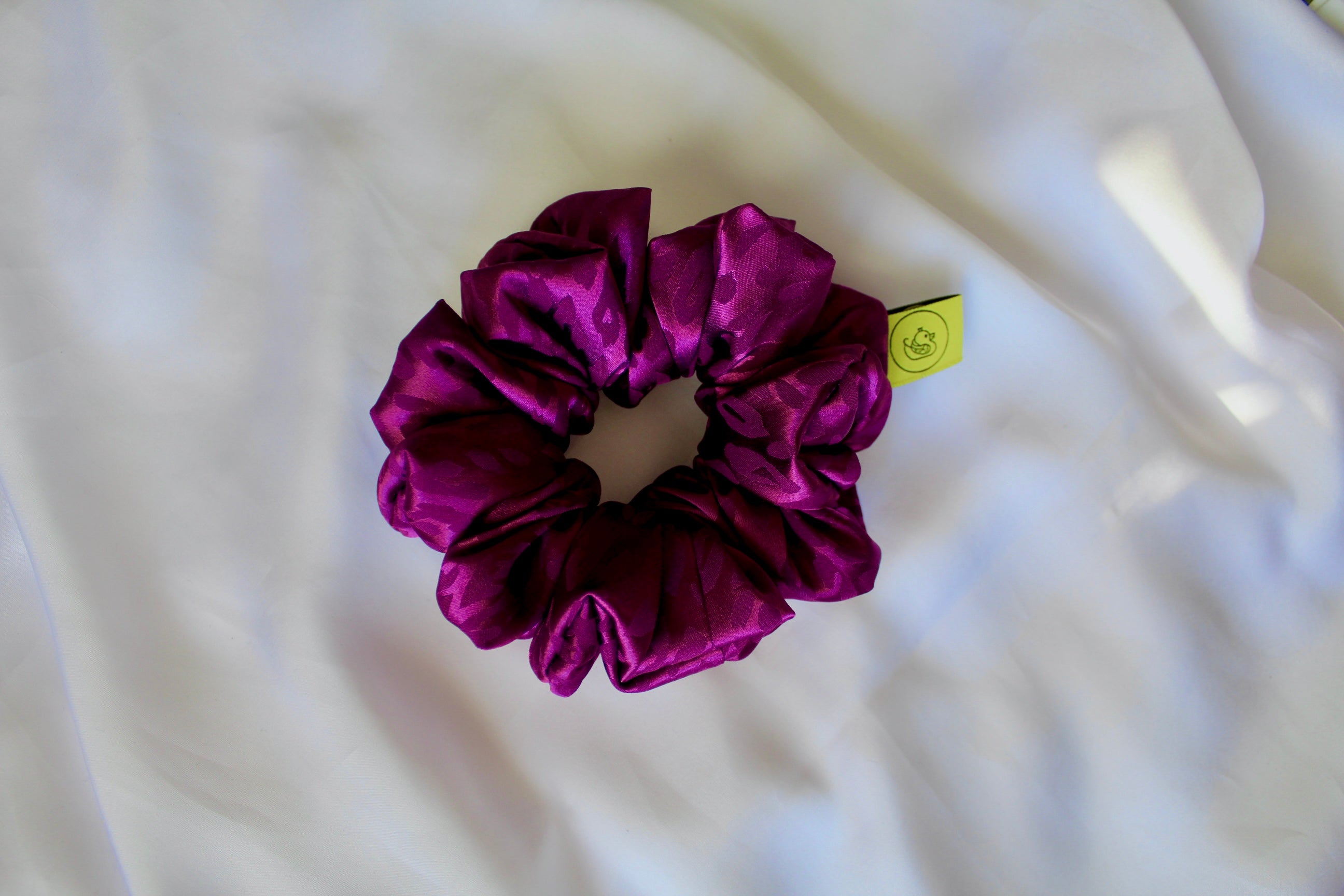 This shows a luxurious scrunchie made of silky textured fabric in a bright deep purple colour.The fabric has a subtle leopard print pattern wove into it, giving it an elegant and slightly reflective appearance.The scrunchie is gathered and voluminous creating soft folds and a plush look.A small yellow label tag with a picture of a duck is attached to the scrunchie adding a touch of branding.The overall aesthetic is soft, elegant and stylish. Perfect for those with sensitive scalp or hair loss