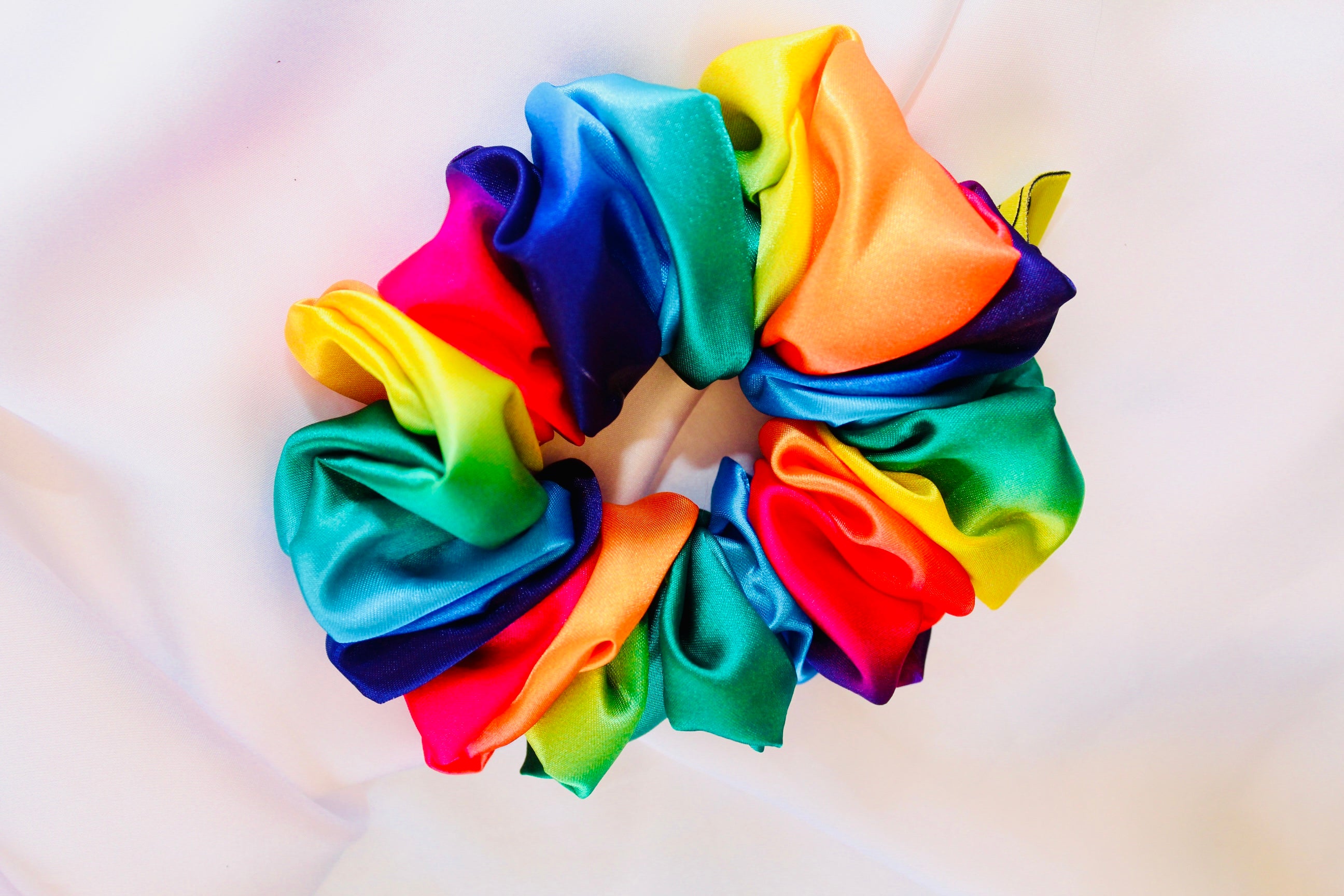 This shows a luxurious scrunchie made of silky textured fabric in a rainbow multi coloured. giving it an elegant and slightly reflective appearance.The scrunchie is gathered and voluminous creating soft folds and a plush look.A small yellow label tag with a picture of a duck is attached to the scrunchie adding a touch of branding.The overall aesthetic is soft, elegant and stylish. Perfect for those with sensitive scalp or hair loss