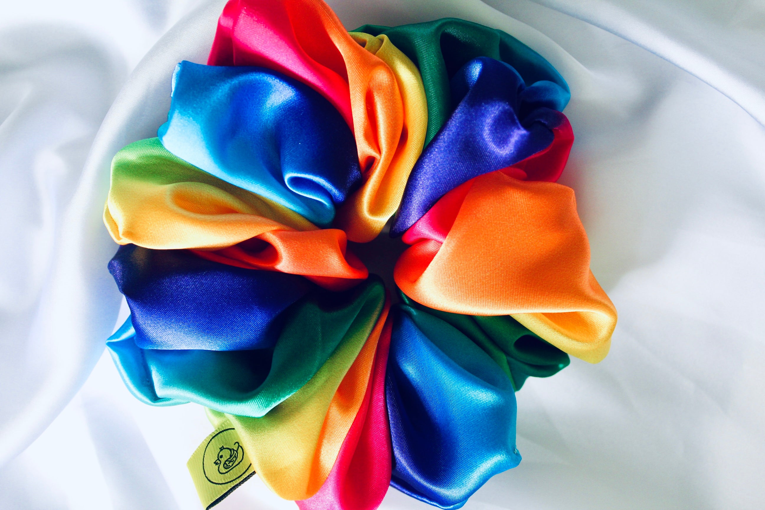 This shows a luxurious scrunchie made of silky textured fabric in a multi colour rainbow running through the fabric giving it an elegant and slightly reflective appearance.The scrunchie is gathered and voluminous creating soft folds and a plush look.A small yellow label tag with a picture of a duck is attached to the scrunchie adding a touch of branding.The overall aesthetic is soft, elegant and stylish. Perfect for those with sensitive scalp or hair loss