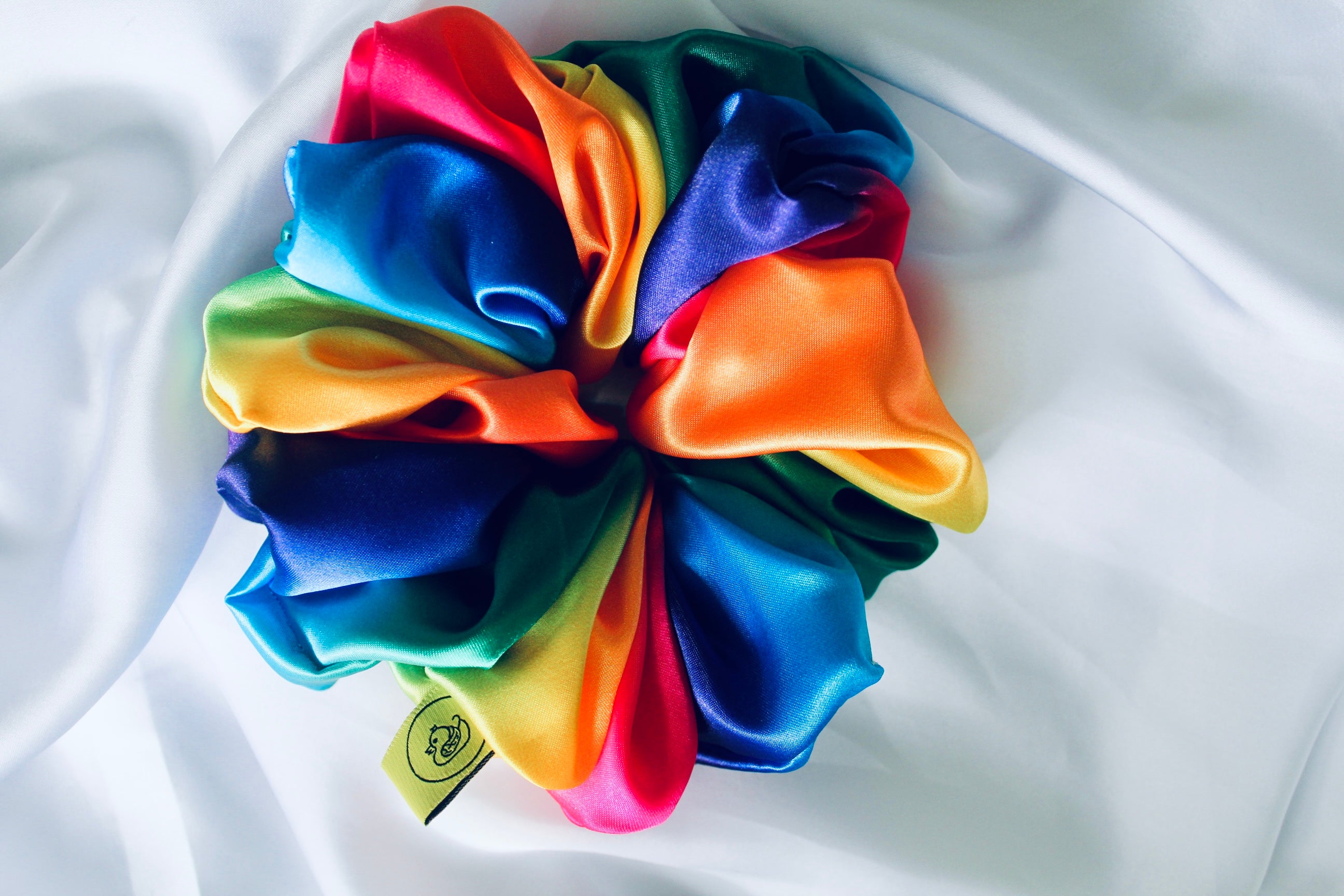 This shows a luxurious scrunchie made of silky textured fabric in a multi colour rainbow running through the fabric giving it an elegant and slightly reflective appearance.The scrunchie is gathered and voluminous creating soft folds and a plush look.A small yellow label tag with a picture of a duck is attached to the scrunchie adding a touch of branding.The overall aesthetic is soft, elegant and stylish. Perfect for those with sensitive scalp or hair loss