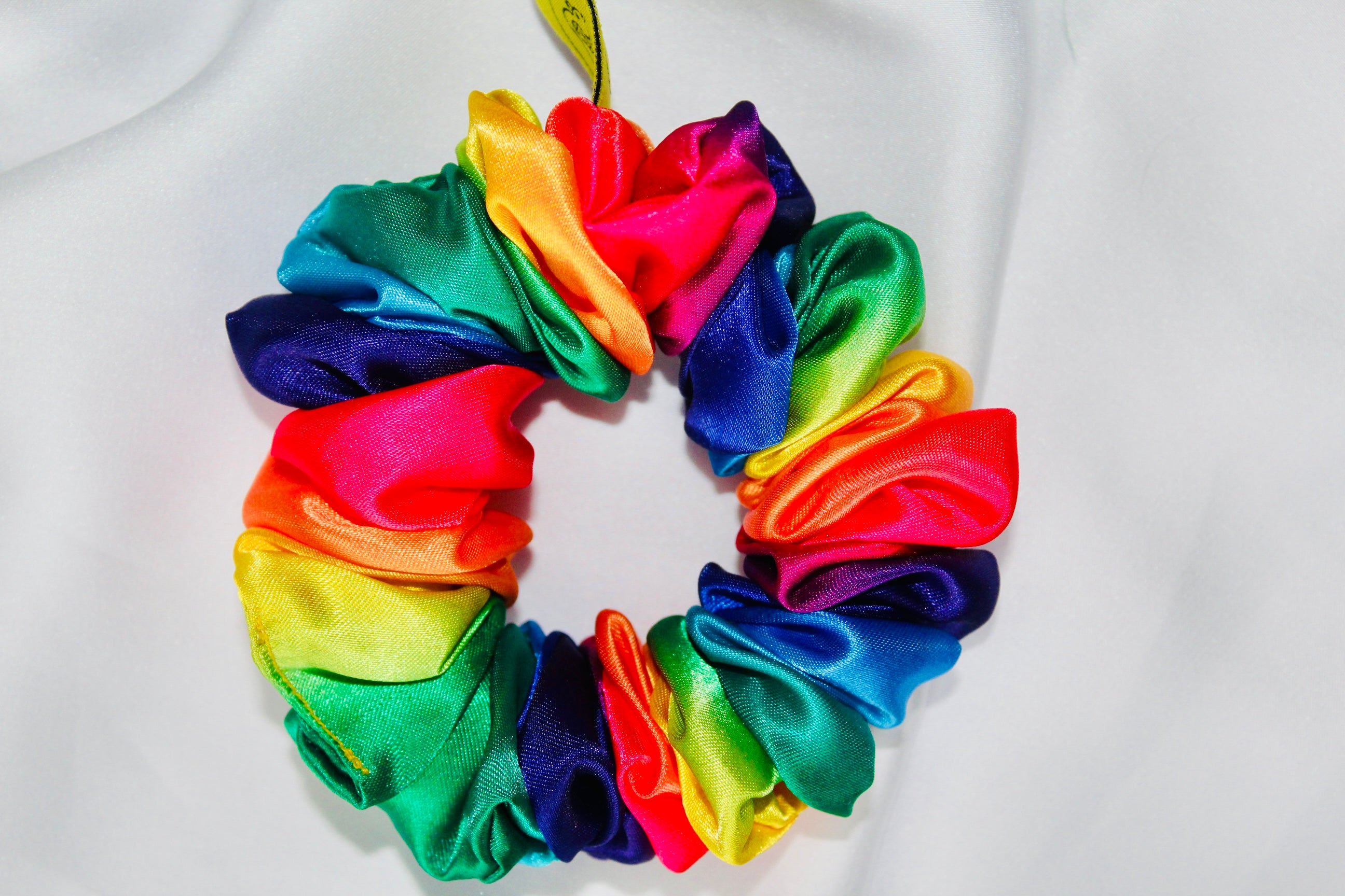 This shows a luxurious scrunchie made of silky textured fabric in a multi colour rainbow. The fabric has a subtle floral pattern, giving it an elegant and slightly reflective appearance.The scrunchie is gathered and voluminous creating soft folds and a plush look.A small yellow label tag with a picture of a duck is attached to the scrunchie adding a touch of branding.The overall aesthetic is soft, elegant and stylish. Perfect for those with sensitive scalp or hair loss