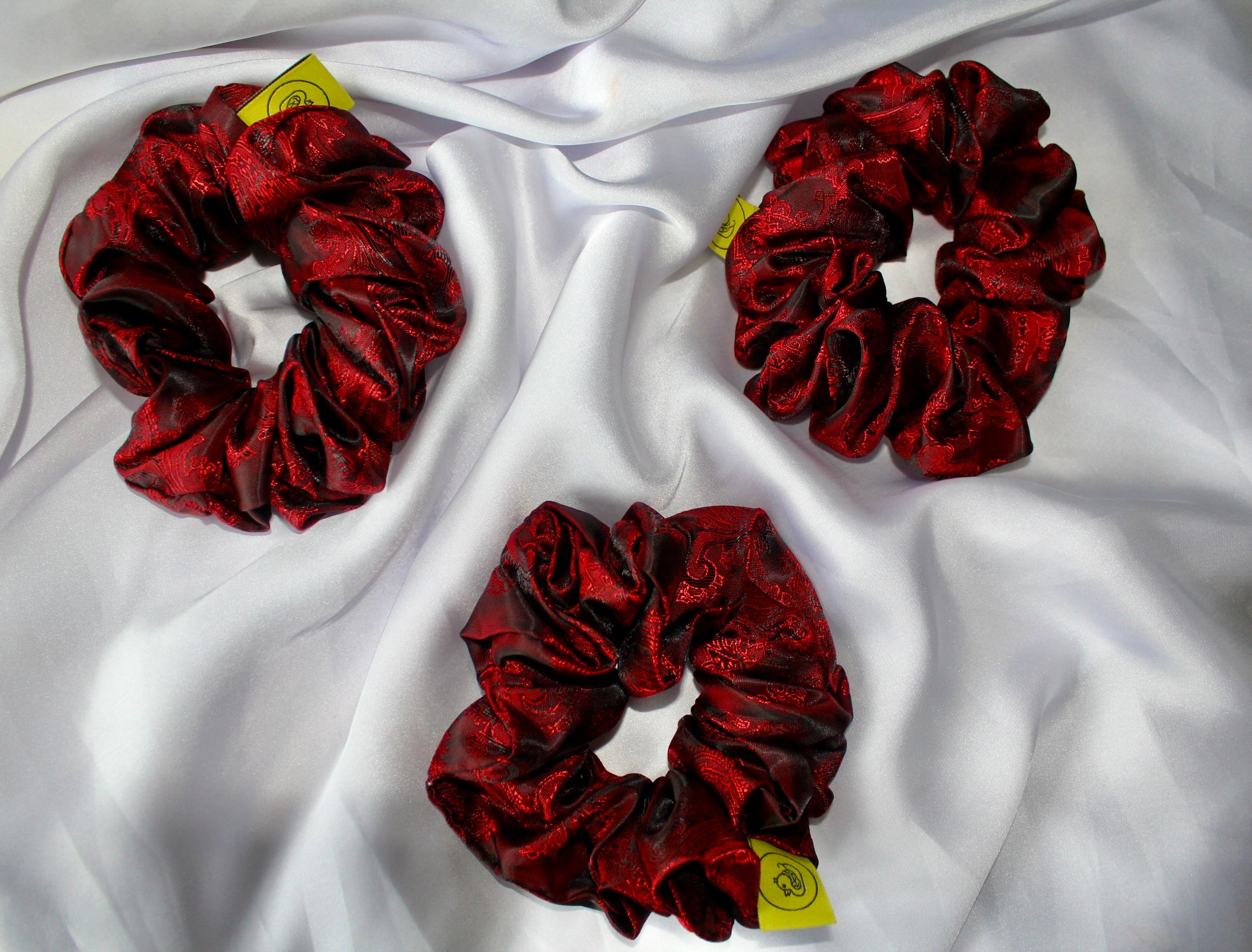 This shows a luxurious scrunchie made of silky textured fabric in a red and black colour.The fabric has a subtle paisley pattern wove into it, giving it an elegant and slightly reflective appearance.The scrunchie is gathered and voluminous creating soft folds and a plush look.A small yellow label tag with a picture of a duck is attached to the scrunchie adding a touch of branding.The overall aesthetic is soft, elegant and stylish. Perfect for those with sensitive scalp or hair loss