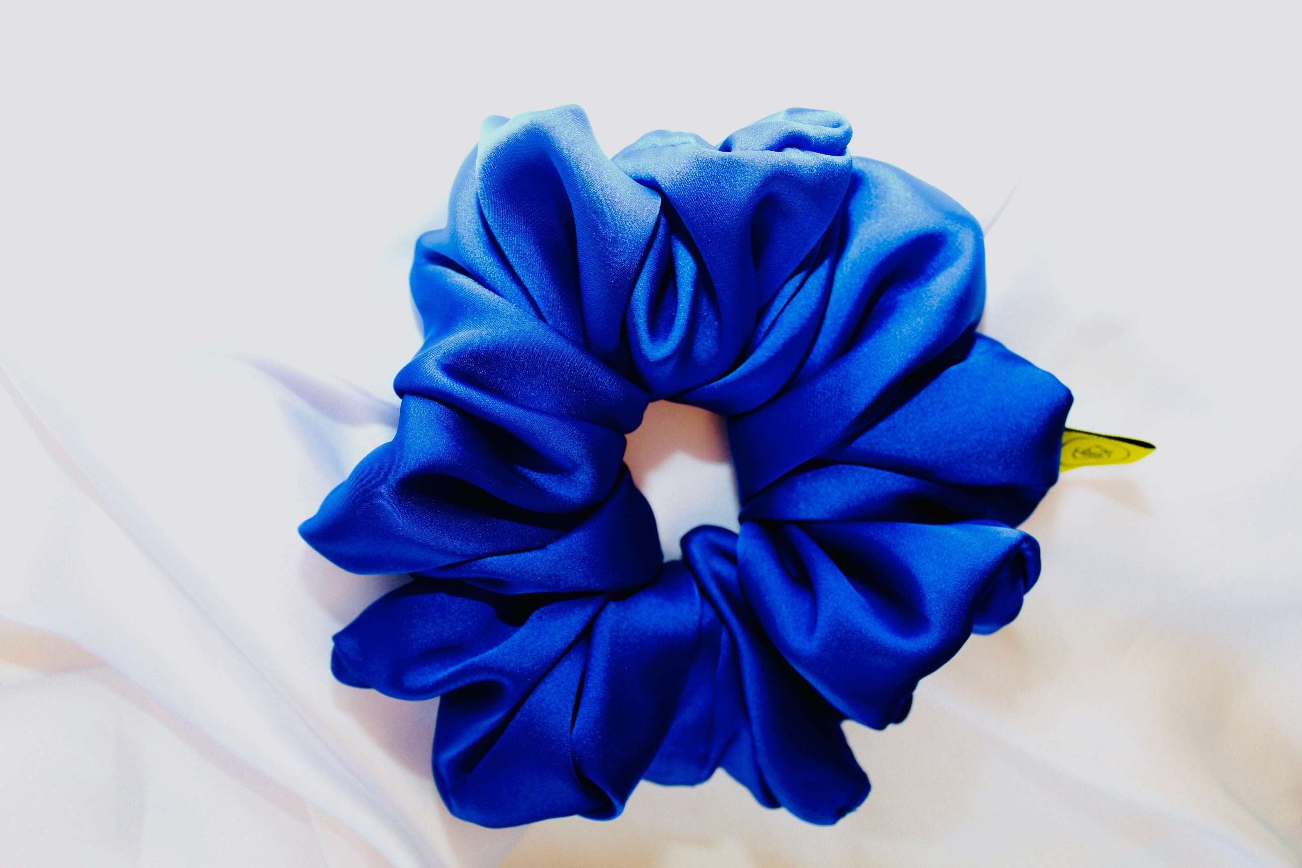 This shows a luxurious scrunchie made of silky textured fabric in a Royal Blue giving it an elegant and slightly reflective appearance.The scrunchie is gathered and voluminous creating soft folds and a plush look.A small yellow label tag with a picture of a duck is attached to the scrunchie adding a touch of branding.The overall aesthetic is soft, elegant and stylish. Perfect for those with sensitive scalp or hair loss