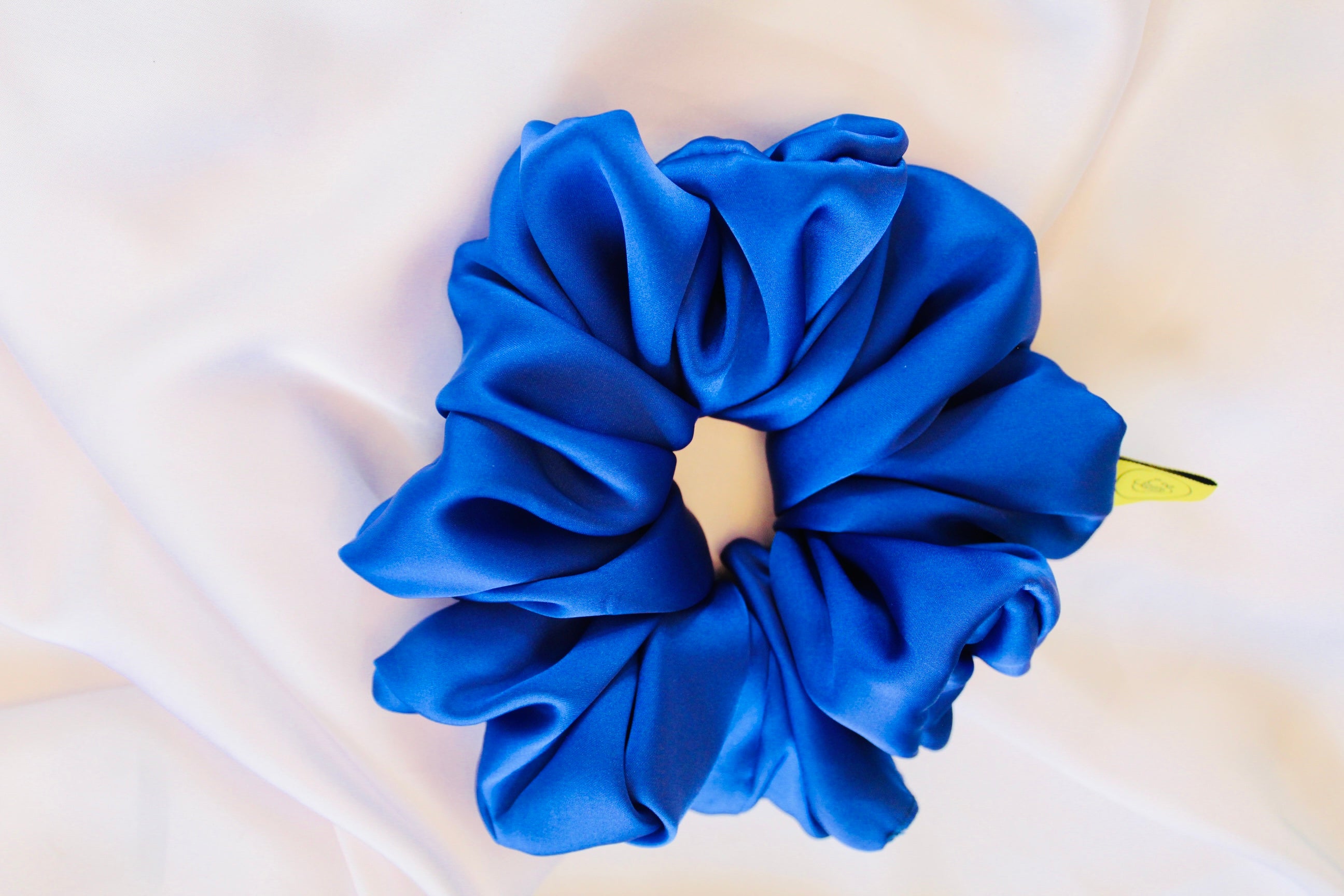 This shows a luxurious scrunchie made of silky textured fabric in a Royal Blue giving it an elegant and slightly reflective appearance.The scrunchie is gathered and voluminous creating soft folds and a plush look.A small yellow label tag with a picture of a duck is attached to the scrunchie adding a touch of branding.The overall aesthetic is soft, elegant and stylish. Perfect for those with sensitive scalp or hair loss
