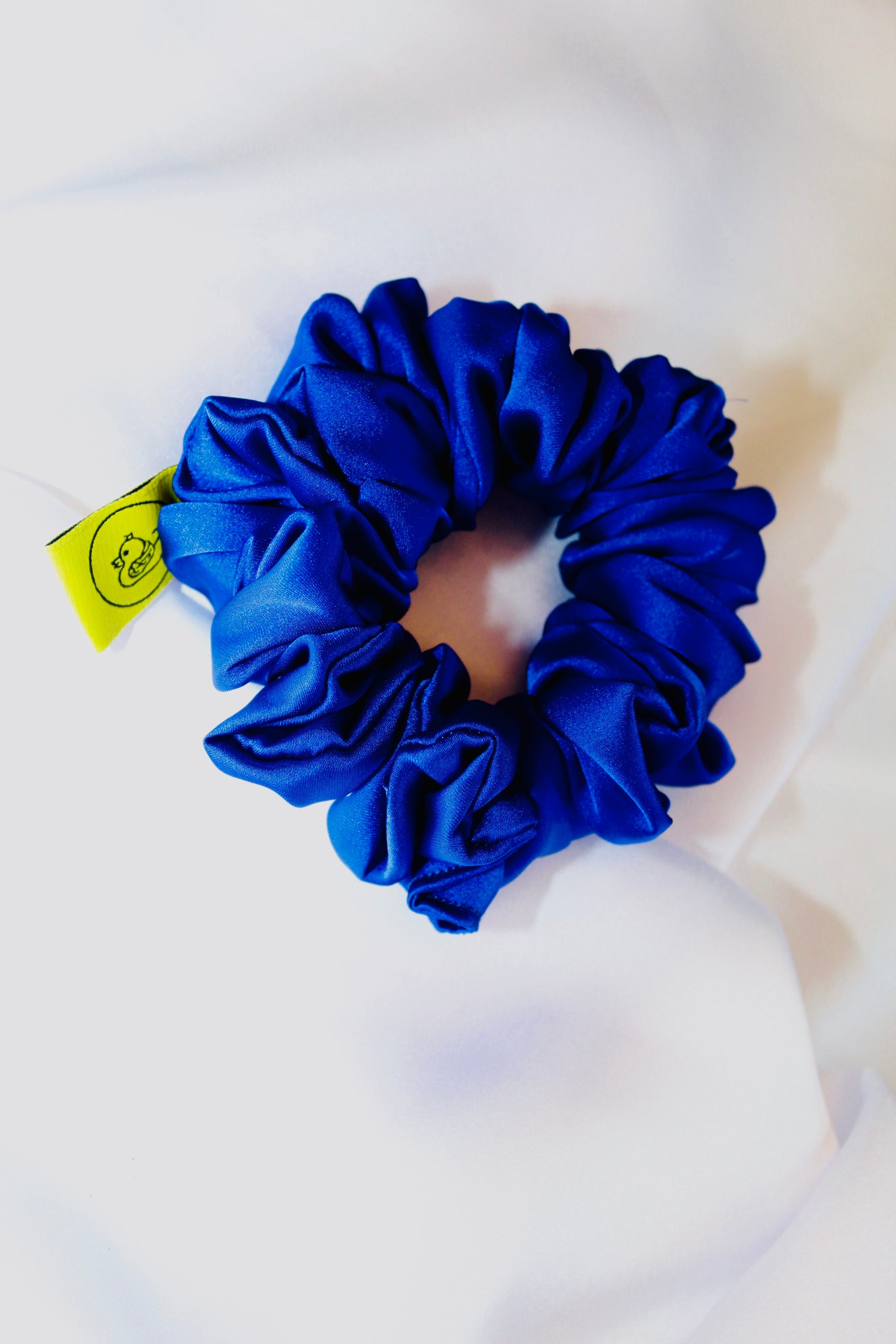This shows a luxurious scrunchie made of silky textured fabric in an Royal Blue. The fabric has a subtle floral pattern, giving it an elegant and slightly reflective appearance.The scrunchie is gathered and voluminous creating soft folds and a plush look.A small yellow label tag with a picture of a duck is attached to the scrunchie adding a touch of branding.The overall aesthetic is soft, elegant and stylish. Perfect for those with sensitive scalp or hair loss