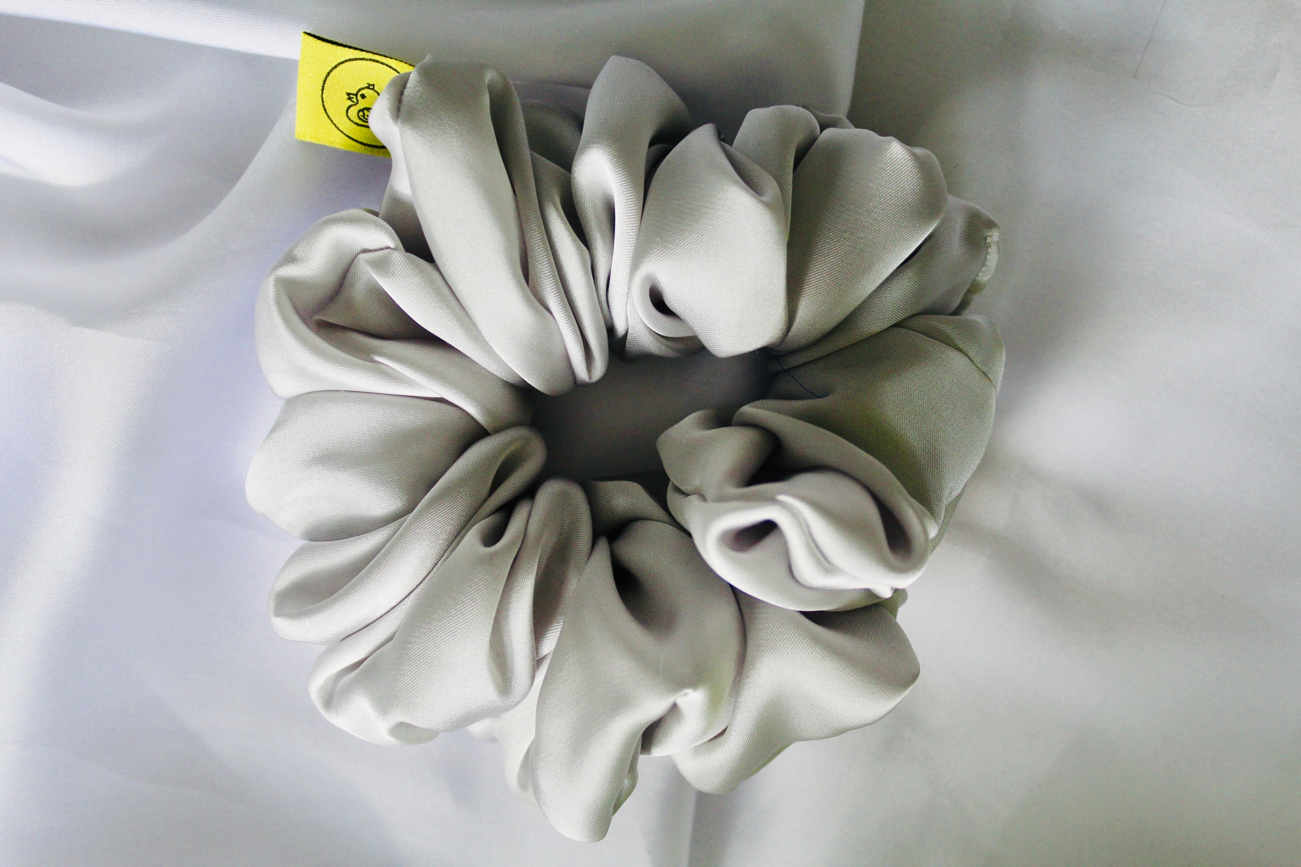 This shows a luxurious scrunchie made of silky textured fabric in a Silver giving it an elegant and slightly reflective appearance.The scrunchie is gathered and voluminous creating soft folds and a plush look.A small yellow label tag with a picture of a duck is attached to the scrunchie adding a touch of branding.The overall aesthetic is soft, elegant and stylish. Perfect for those with sensitive scalp or hair loss