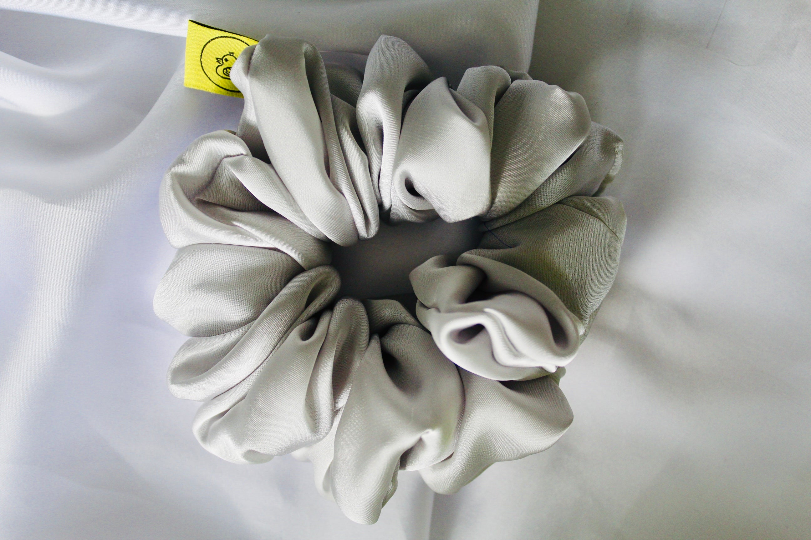 This shows a luxurious scrunchie made of silky textured fabric in a Silver giving it an elegant and slightly reflective appearance.The scrunchie is gathered and voluminous creating soft folds and a plush look.A small yellow label tag with a picture of a duck is attached to the scrunchie adding a touch of branding.The overall aesthetic is soft, elegant and stylish. Perfect for those with sensitive scalp or hair loss